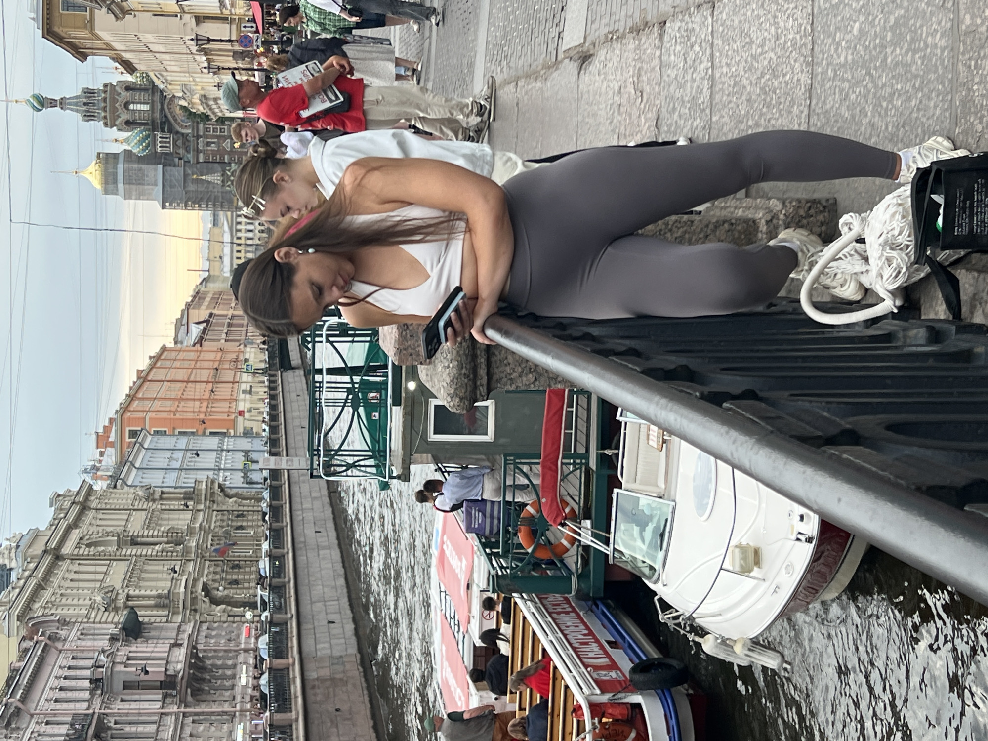 The image depicts a daytime scene in Saint Petersburg, Russia, with a canal in the foreground. Several boats, including tour boats, are docked or moving on the water. In the background, ornate buildings line the canal, and a prominent onion-domed church is visible.
In the immediate foreground, a young woman in grey leggings and a white tank top is leaning on a railing, looking down at her phone. Another young woman in a white top and pink headband is partially visible behind her. The railing is made of dark metal with a decorative design. To the right, a white boat with red trim is docked, and a white mop with a white rope is resting on the ground next to it.
Further down the canal, more people are seen on other boats, some appearing to be passengers on tours. The architecture of the buildings suggests a European city, and the specific style of the church and the canal setting are consistent with Saint Petersburg. The lighting suggests it is daytime, possibly late afternoon given the soft light. There are no overt emotional expressions visible on the faces of the people in focus, but the overall atmosphere appears casual and urban. Some text is visible on a sign on one of the tour boats, but it is not legible at this resolution.