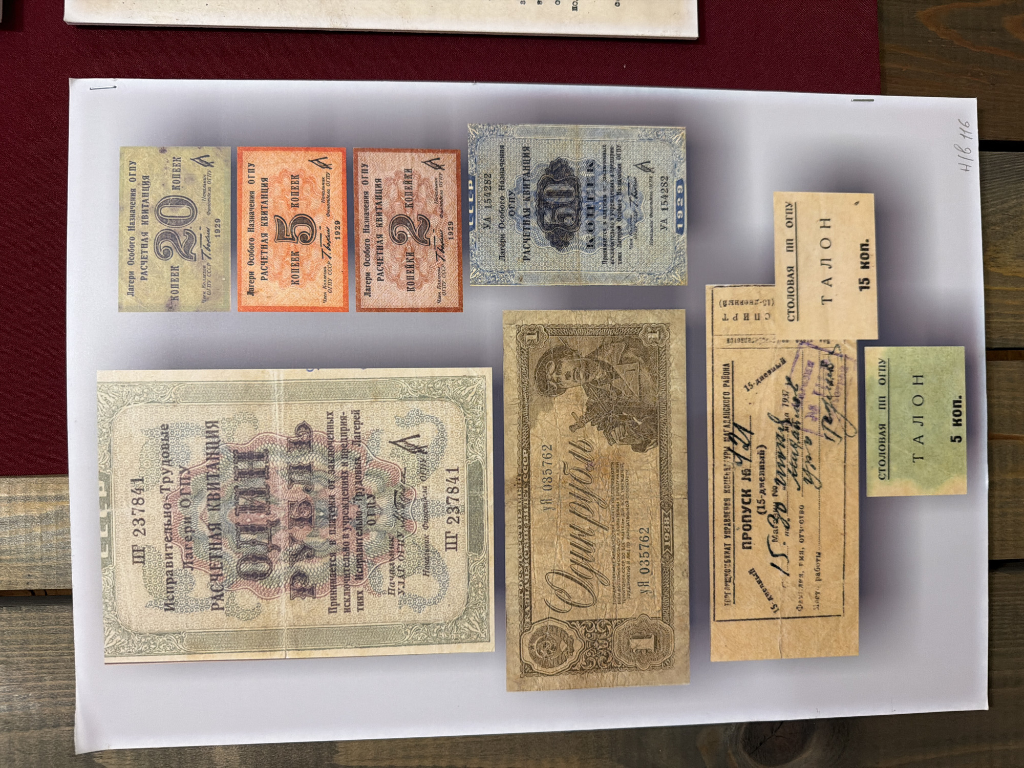 The image displays a collection of historical Soviet documents, primarily financial and ration coupons, arranged on a white surface with a wooden background. The documents include:
A large banknote with the denomination "ОДИН РУБЛЬ" (One Ruble) and the text "Расчетная квитанция" (Settlement Receipt). It bears the stamp "ШГ 237841".
Several smaller coupons with denominations in "КОПЕЕК" (Kopecks), including "20 КОПЕЕК," "5 КОПЕЕК," and "50 КОПЕЕК." These also state "Расчетная квитанция" and "Лагеря Особого Назначения ОГПУ" (Camps of Special Purpose OGPU), with dates from 1929.
A coupon with "ОДИН РУБЛЬ" (One Ruble) and the inscription "УЯ 035762," dated 1933.
A "ПРОПУСК № 141" (Pass No. 141) dated "15-дневный" (15-day) and "1937," issued by "УПРАВЛЕНИЯ КОМБИ NAT УРАЛГАНСКОГО РАЙОНА" (Management of Kombinat Uralsky District). It includes handwritten entries for names and possibly work assignments.
Two ration coupons labeled "ТАЛОН" (Coupon), one for "5 коп." and another for "15 коп."
There is no discernible information within the image that directly or indirectly references the city of Ust'-Pyzas, Russia.