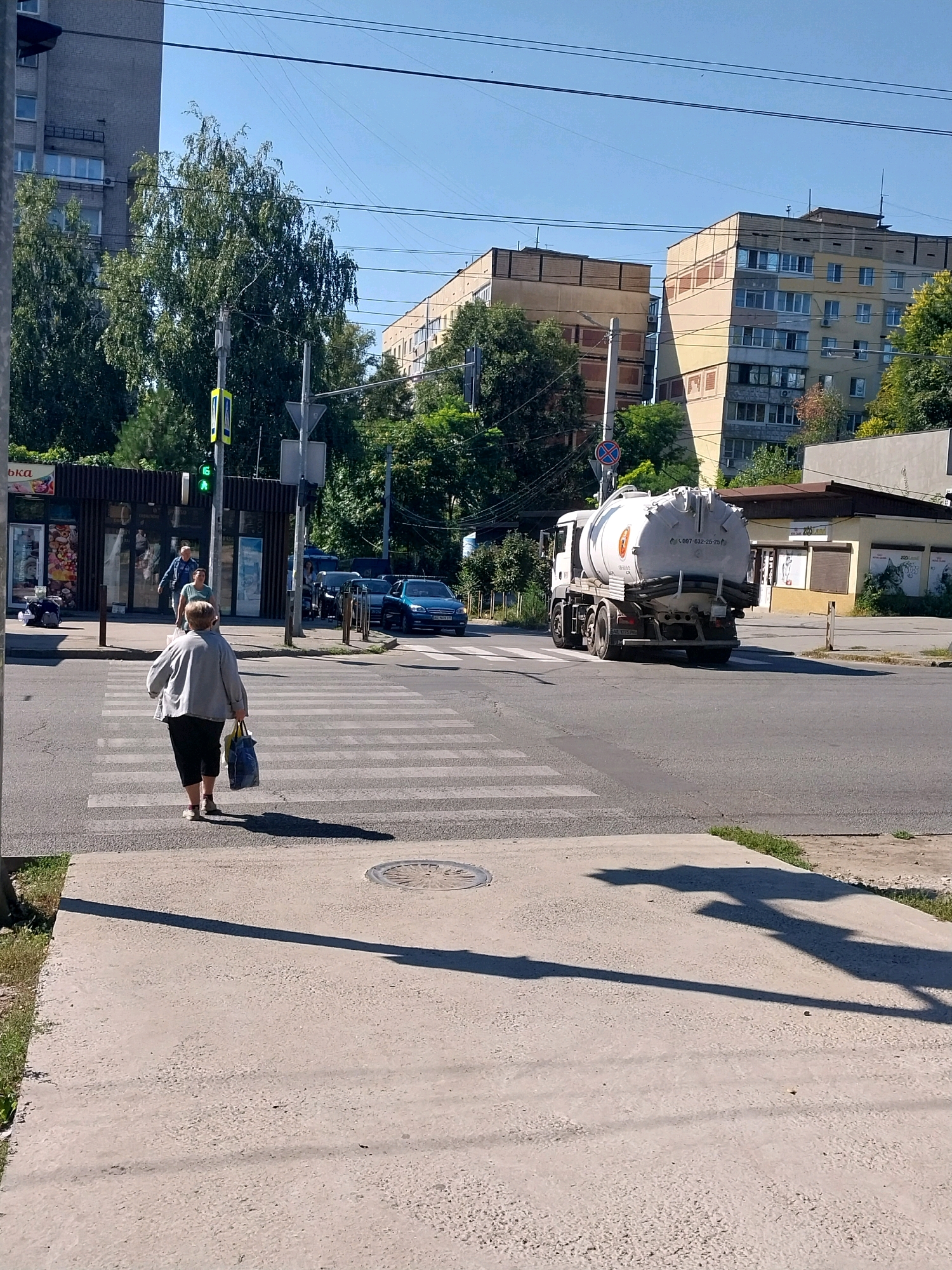 The image depicts a street scene in Dnipro, Ukraine, on a sunny day. In the foreground, a pedestrian crossing is visible with a woman walking across it, carrying two shopping bags. Behind her, a large white tanker truck with the license plate "AZ 1775 PM" is driving across the intersection. In the background, several buildings, trees, and traffic lights are present. The buildings appear to be apartment complexes and commercial establishments. The overall environment suggests an urban setting.