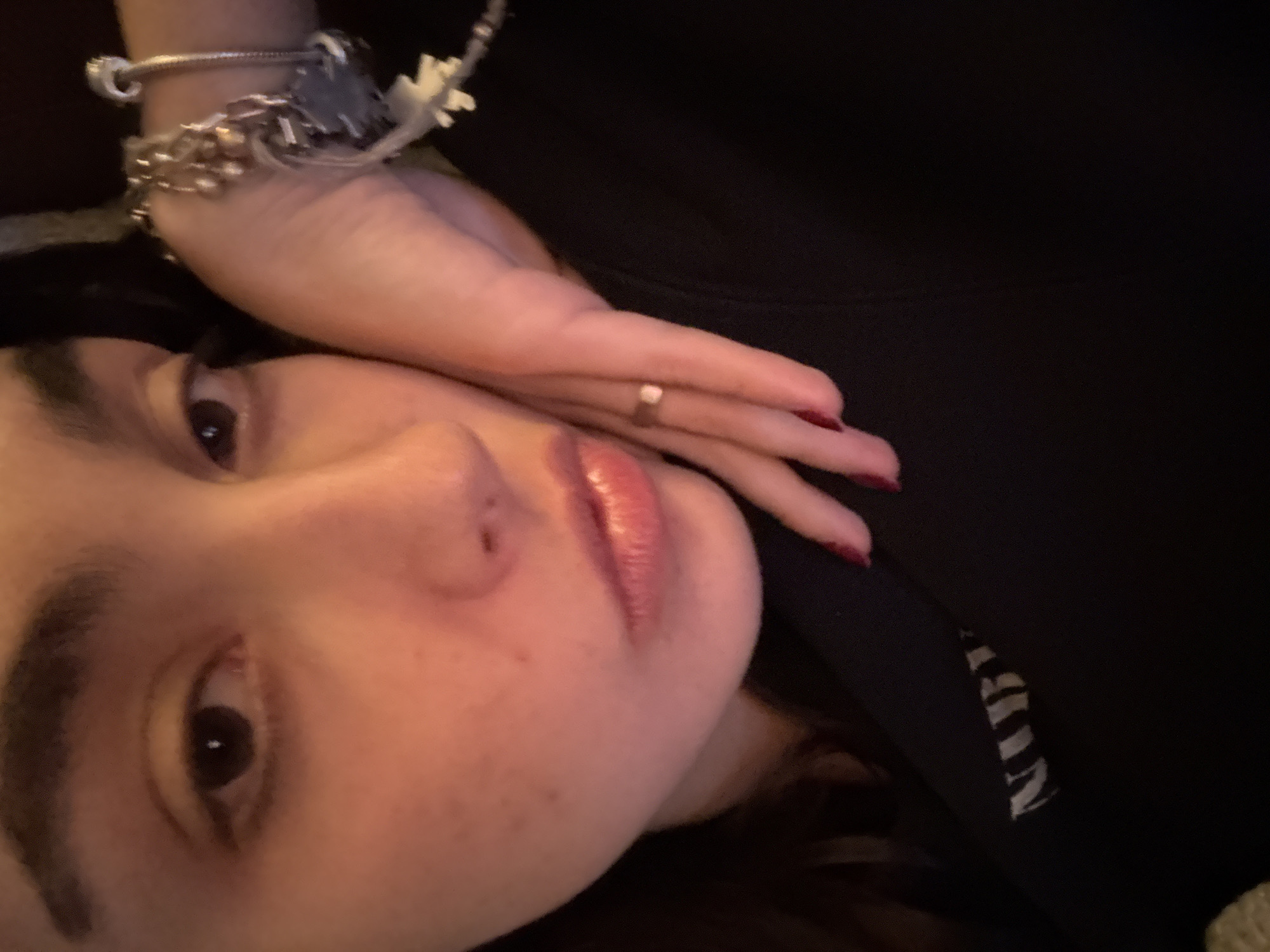 A close-up, angled self-portrait of a woman's face and upper chest. She has fair skin, dark hair, and brown eyes. Her left hand is positioned near her face, adorned with a silver charm bracelet and a simple ring on one of her fingers. The woman is wearing a dark-colored garment with white lettering visible along the collar. The location of the scene is not indicated.