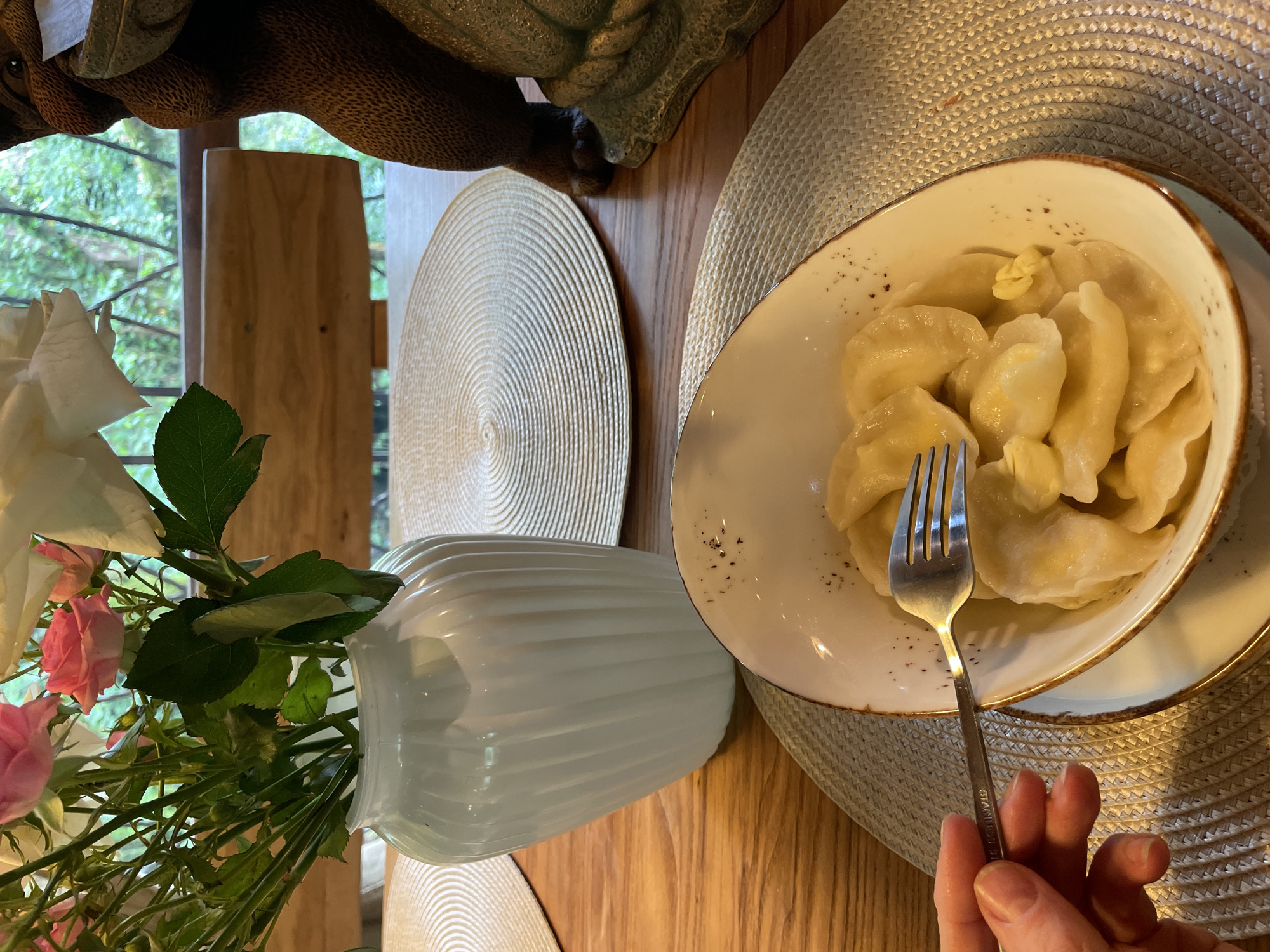 The image shows a dining table scene, likely indoors. A white vase with roses and greenery sits on the table, adjacent to a tan woven placemat. Another similar placemat is visible in the background. A bowl of pierogi, topped with butter, is on a plate atop one of the placemats. A hand is holding a fork, presumably about to eat the pierogi. The table is made of wood. Through a nearby window, greenery is visible, potentially suggesting a rural or garden setting. This image was likely taken in Solokh-Aul, Russia.