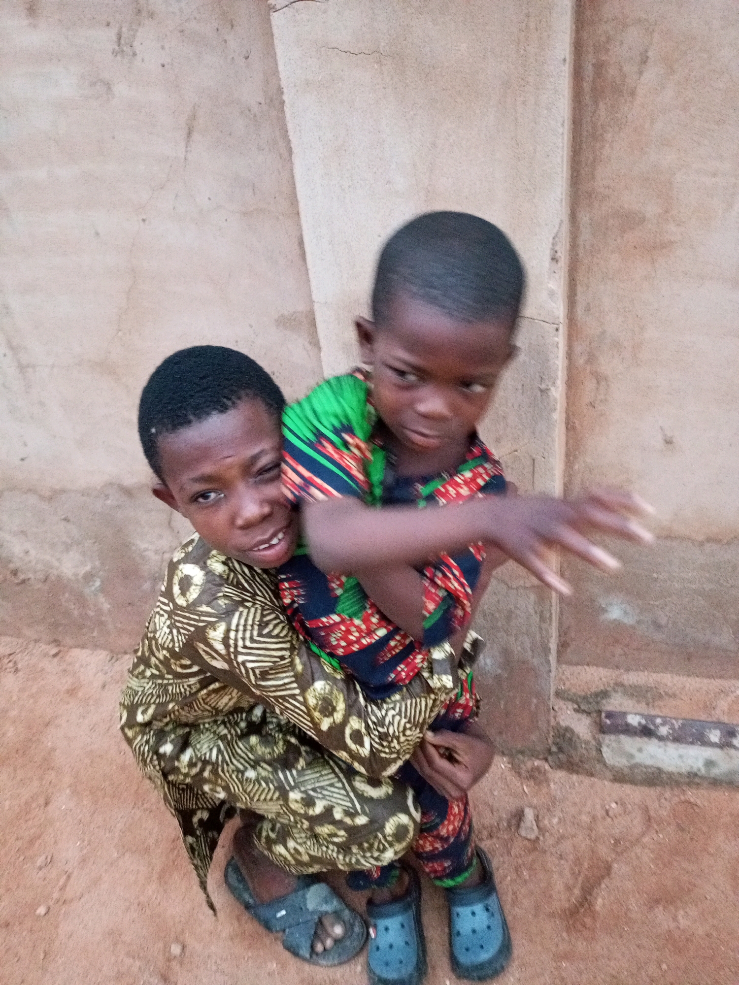 Two young Black children are posing for a photo outside a textured, pale wall. The child in the foreground, on the left, is wearing a brown and white patterned garment and dark sandals. The child behind, on the right, is wearing a green, red, and black patterned shirt and dark blue crocs. The child in front appears to be looking at the camera with a slightly crooked smile. The child behind is looking away from the camera, with their right hand extended. The ground beneath them is sandy and reddish-brown, with small pebbles and debris. The wall behind them is a light beige color with visible imperfections, cracks, and a protruding horizontal element near the bottom. The lighting suggests it is daytime. The context of Tempure, Nigeria, implies this is an outdoor scene in a likely rural or semi-urban setting.