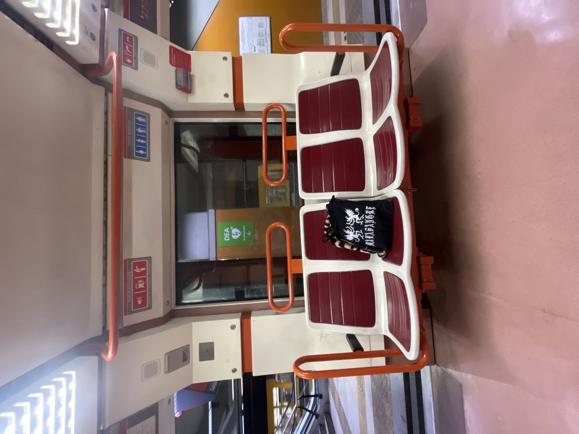 A photograph taken indoors depicts an interior view of a commuter train carriage in Buenos Aires, Argentina. The carriage features two sets of facing seats upholstered in maroon with white frames and orange handrails. On one seat is a black tote bag with white writing and tiger print accents. Safety signage and evacuation plans are visible on the walls of the carriage. The floor in the train carriage is pink, and a window provides a view of the outdoor environment.