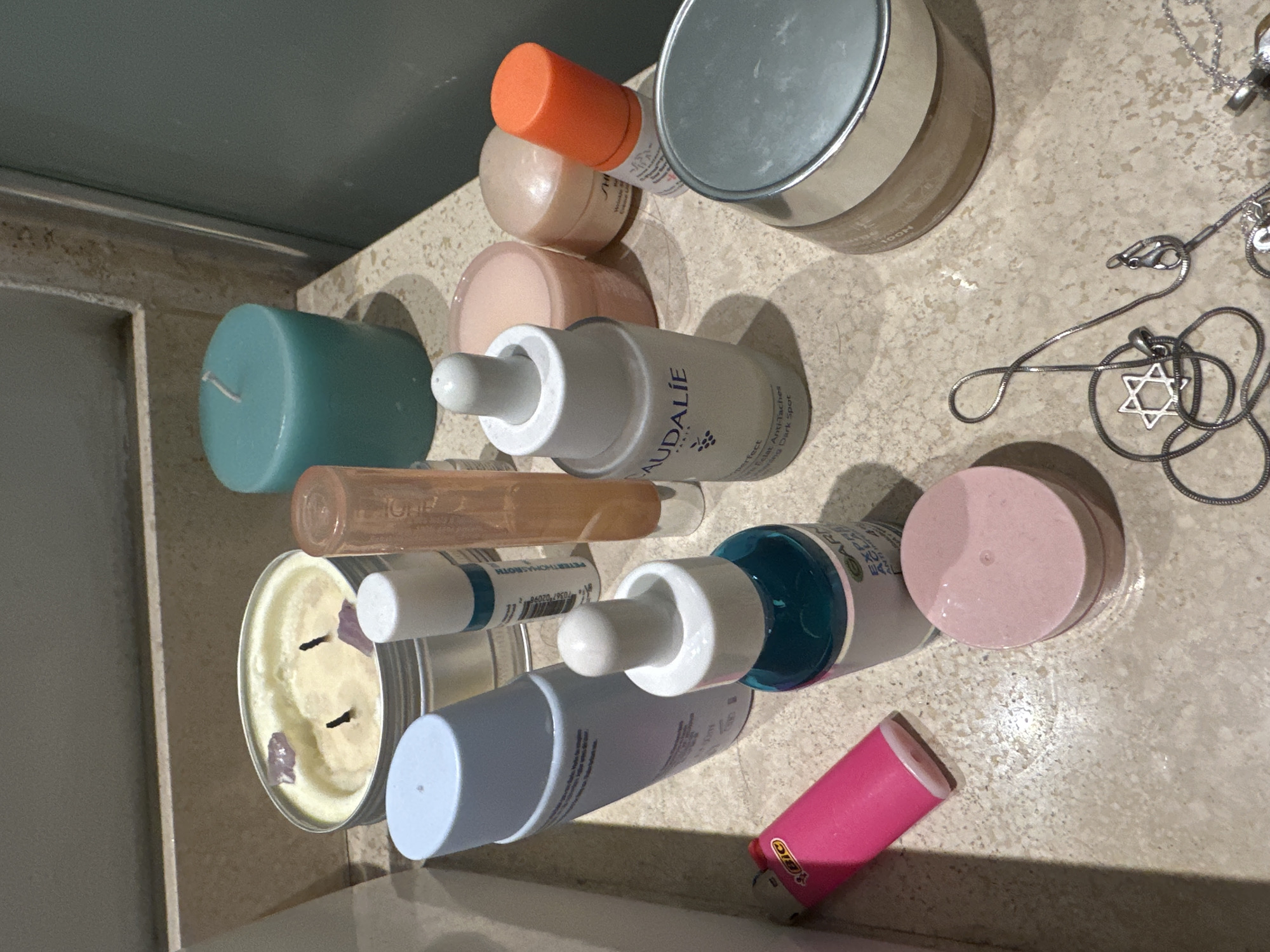 A collection of personal care items, candles, and jewelry is arranged on a speckled, light-colored countertop, likely in a bathroom or dressing area, within a residence in Huixquilucan de Degollado, Mexico.
The subjects are various objects including skincare products, a lighter, candles, and necklaces. No people are visible. The scene captures a static arrangement of these items, suggesting daily use or storage, with no specific activity taking place.
Notable details include a large, silver-lidded jar (possibly a cream), several dropper bottles for serums or oils, and smaller jars and bottles. A prominent teal cylindrical candle and a wider, pale yellow candle in a metal tin with two wicks and visible crystals (possibly amethyst) are present. A pink BIC lighter is also visible. Jewelry, including several silver-colored necklaces, one featuring a Star of David pendant and another with a heart charm, are scattered on the counter. The lighting is artificial and indoor, providing no clues about the time of day or weather.
Visible text on the product labels includes:
* "CAUDALIE"
* "Vinoperfect"
* "Sérum Anti-Taches"
* "Radiance Serum Anti-Dark Spot"
* "PETER THOMASROTH"
* "Wrinkle" (partially visible)
* "BIC" (on the lighter)
* "0367 02096" (on a product label)
* Partial brand names that appear to be "Drunk Elephant" (from an orange-capped bottle) and "TOOH" (on another container).