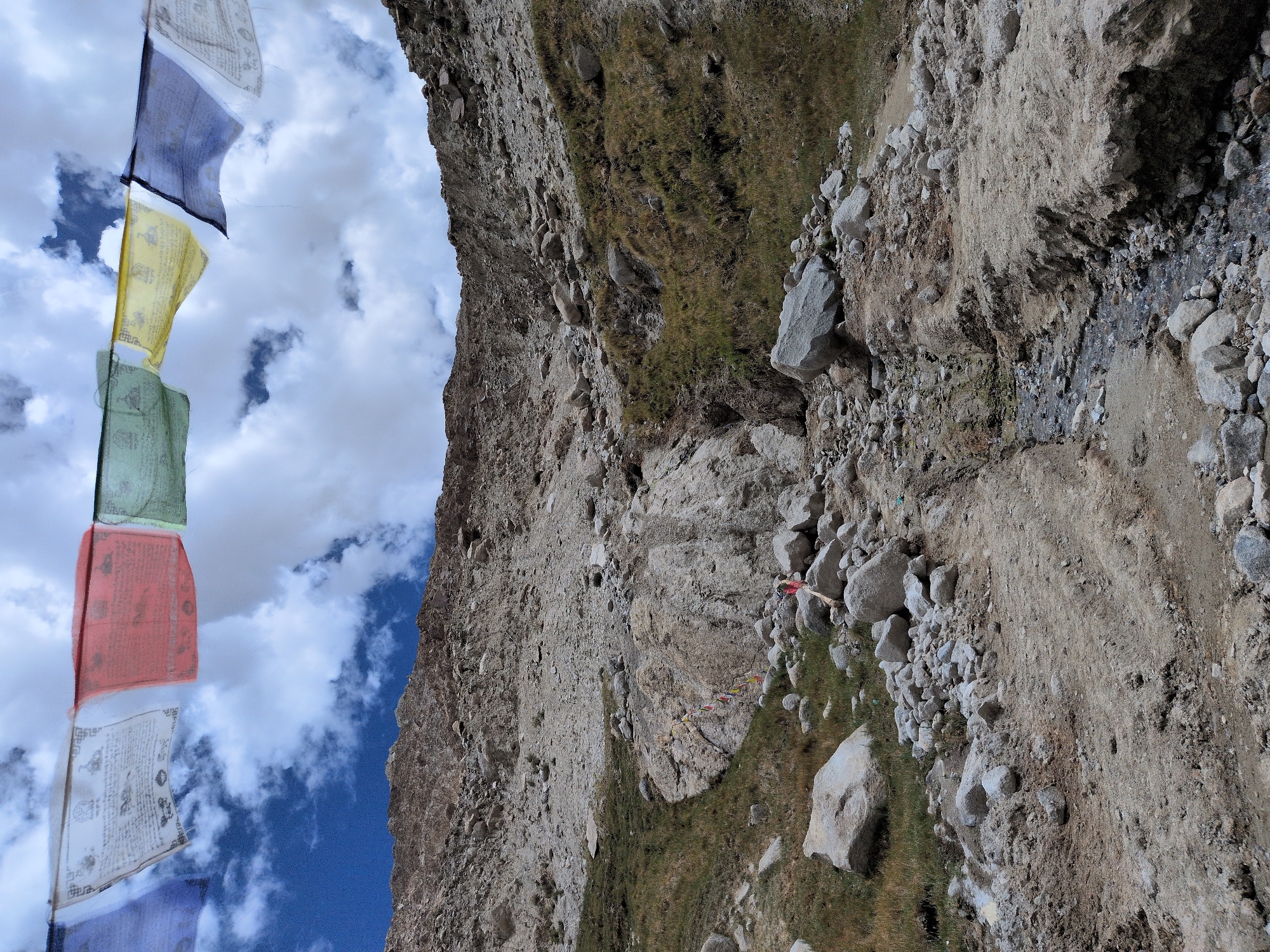 The image depicts a rugged, rocky mountain slope in Panamik, India, under a cloudy sky. Prayer flags, displaying various colors including blue, white, red, green, and yellow, are strung across the upper portion of the frame, fluttering in the wind. The mountainside is covered in loose stones, scree, and patches of dry grass, suggesting a high-altitude, arid environment. A narrow, shallow stream or dry creek bed winds its way down the slope, marked by scattered rocks and a faint track of disturbed earth. In the middle ground, a small cluster of prayer flags are tied to a rock face, adding a splash of color against the natural terrain. The overall impression is one of a remote, windswept landscape characteristic of mountainous regions.