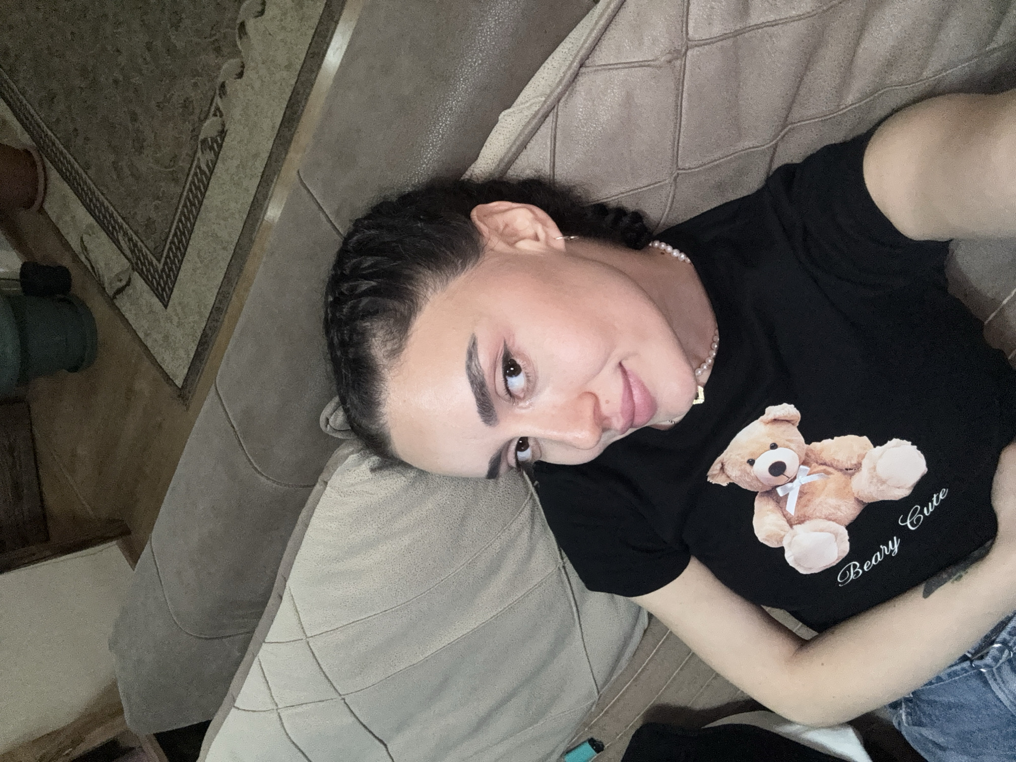 A person is lying on a sofa. The sofa is tan-colored and textured with stitched squares. The person is wearing a black T-shirt with a teddy bear graphic labeled "Beary Cute." The person has dark hair styled in braids and is wearing a pearl necklace. Part of a patterned rug and what appears to be the edge of a table are visible in the background. A vape device can be seen near the bottom edge of the sofa. The location is likely a residential interior and there is no direct evidence indicating the specific location of Tbilisi, Georgia.