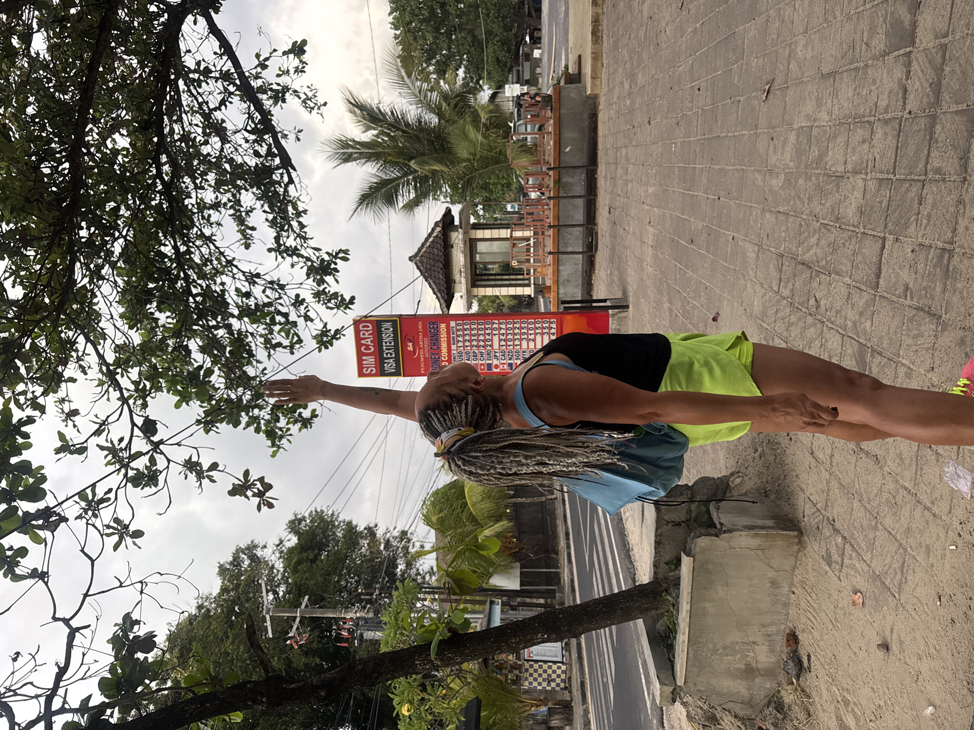 In Kuta, Indonesia, a person with grey braids and a blue backpack stands on a tiled sidewalk, reaching up towards the leaves of a tree. They are wearing a black tank top and bright green shorts. Behind them, a red and white sign displays "SIM CARD VISA EXTENSION" and currency exchange rates. Further in the background, there is a small building resembling a security booth or information kiosk, and palm trees. The street adjacent to the sidewalk is visible, with a car parked in the distance. The sky is overcast, suggesting a cloudy day. The ground on the left side of the image is covered in sand and scattered debris, including cigarette butts.