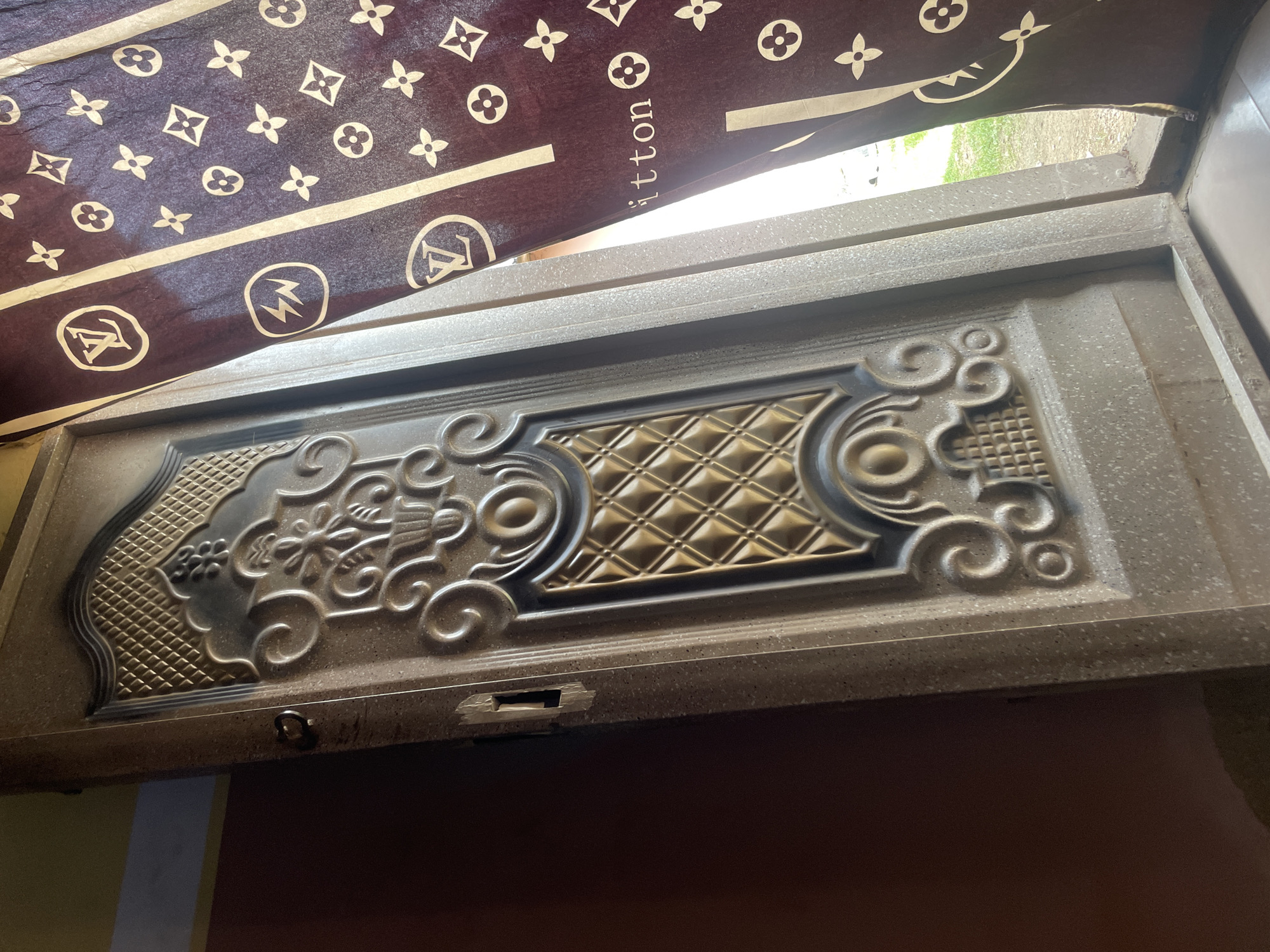 A security door with intricate embossed patterns and a metallic finish stands partially open. To the right of the door, a dark brown curtain with a repeating pattern of white logos and symbols hangs, partially obscuring a view of a bright outdoor area with green grass. The scene is likely located in Jos, Nigeria, a city known for its security consciousness and the prevalence of such ornate doors.