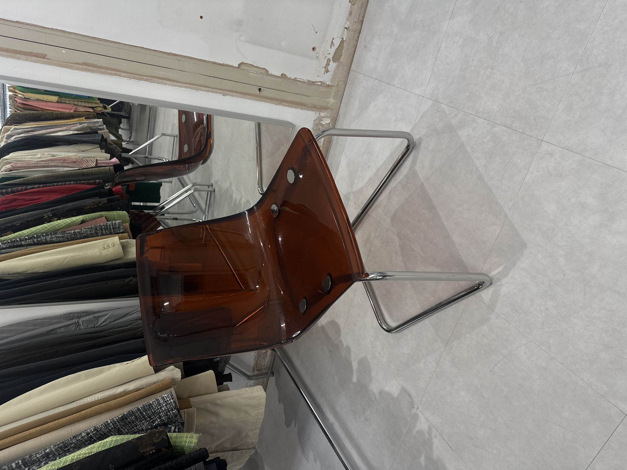 The image depicts a clear acrylic cantilever chair with a brown tint, featuring a sleek, modern design. The chair is placed on a light grey tiled floor. Behind the chair, a rack of clothing is visible, suggesting a retail or storage environment. The clothing includes a variety of garments, such as jackets, coats, and dresses, in different colors and textures. The background also shows a partial view of a mirror, reflecting more clothing racks and a portion of the environment. The overall scene appears to be an indoor setting, possibly a boutique, vintage shop, or a clothing store. The lighting is bright, illuminating the space and casting shadows from the chair and clothing racks. There is no indication of the specific location within London, United Kingdom, nor any people present in the image.