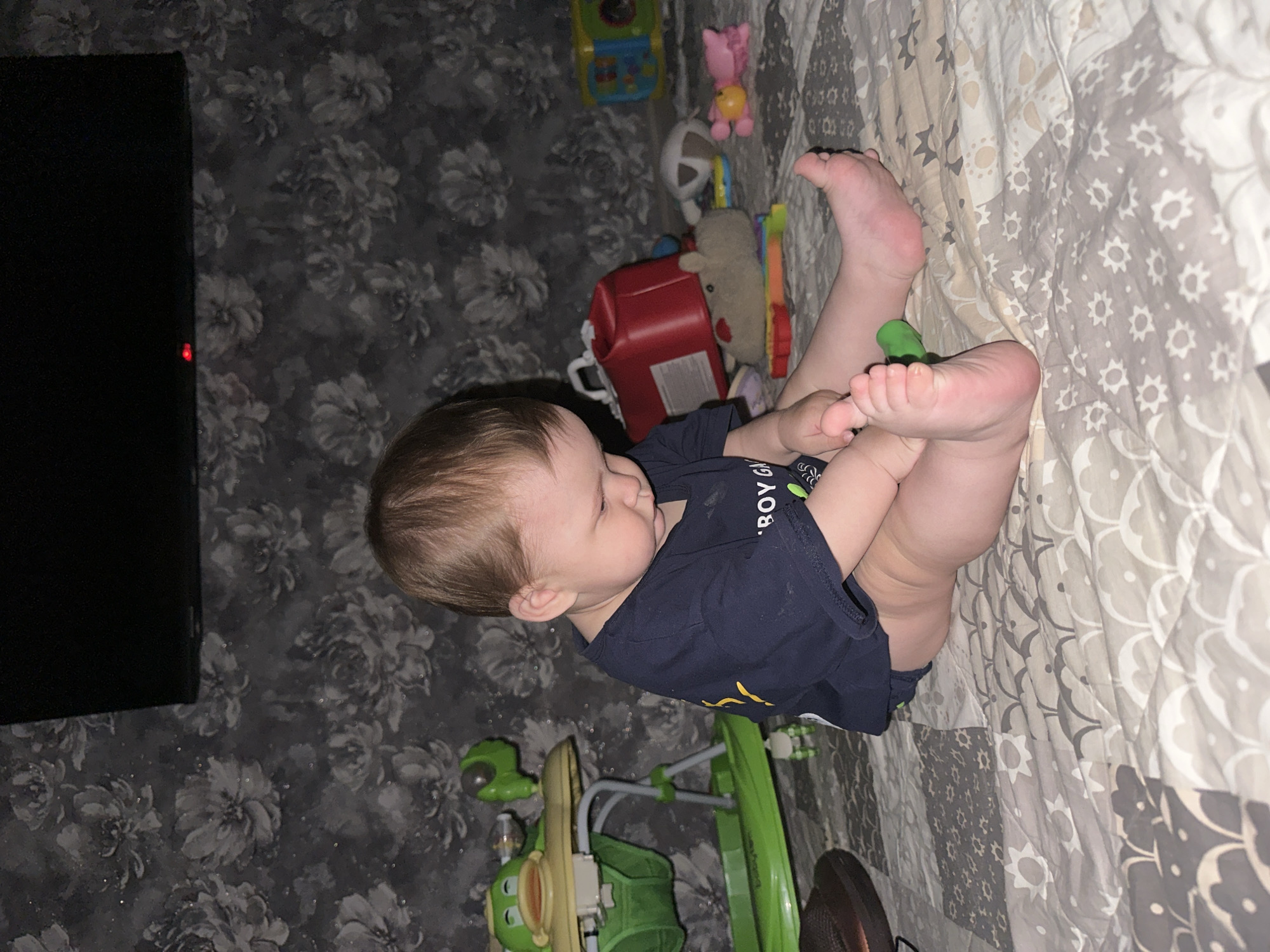 A baby is lying on a patterned blanket with their legs in the air. The baby is wearing a dark blue t-shirt with yellow text on it, and has light brown hair. The baby's feet are visible and appear to be touching a green object. There are various toys scattered around the baby, including a red container, a stuffed animal, and a brightly colored toy. A portion of a black television screen is visible on the left side of the image. The background wallpaper is dark and features a floral pattern. The lighting suggests it could be daytime or evening indoors. The baby's expression is neutral, and the scene appears to be in a domestic setting. The text on the baby's shirt is partially visible and reads "BOY GRL".
