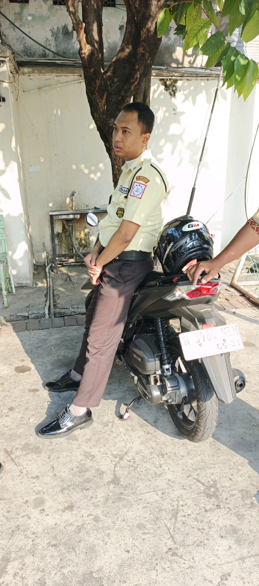 An Indonesian security guard in a light yellow uniform with patches, brown pants, and black dress shoes is sitting on a black motorcycle in front of a building. The guard's hands are clasped in his lap. A black helmet with a "GM" logo is on the motorcycle behind the guard. Another person's hand with a patterned sleeve is visible on the back of the motorcycle. A license plate reading "W 3989 MBH" is attached to the back of the motorcycle. The scene is outdoors, with a concrete ground, a tree, and a wall in the background. The sun is casting shadows.