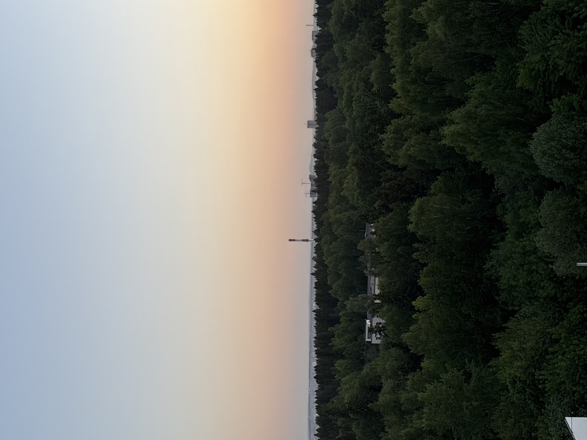 The image depicts a landscape at dusk, seemingly taken from an elevated position. On the left, the sky transitions from a light blue to a pale orange hue. The right side of the image is dominated by a dense forest, the trees' dark green canopy obscuring much of the ground below. A tall, red and white striped chimney or tower is visible in the distance, positioned between the sky and the forest. Interspersed among the trees are glimpses of buildings, suggesting an urban environment bordering the forest. Given the indication that the location is Izhevsk, Russia, the chimney could potentially be identified with further research into industrial structures in that area.