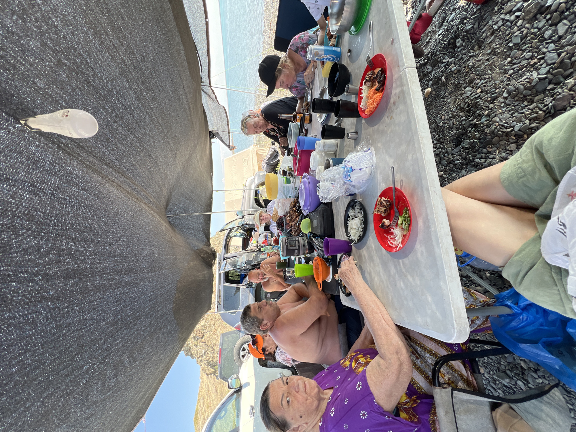 A group of people are gathered around a table for a meal outdoors. The scene appears to be on a rocky shoreline, with the sea visible in the background. Several vehicles are parked nearby, suggesting a camping or picnic outing. A large tarp provides shade over the gathering. Food and drinks are spread across the table, and some individuals are actively eating. The overall atmosphere is casual and social. The location is identified as the city of Karashygan, Kazakhstan.