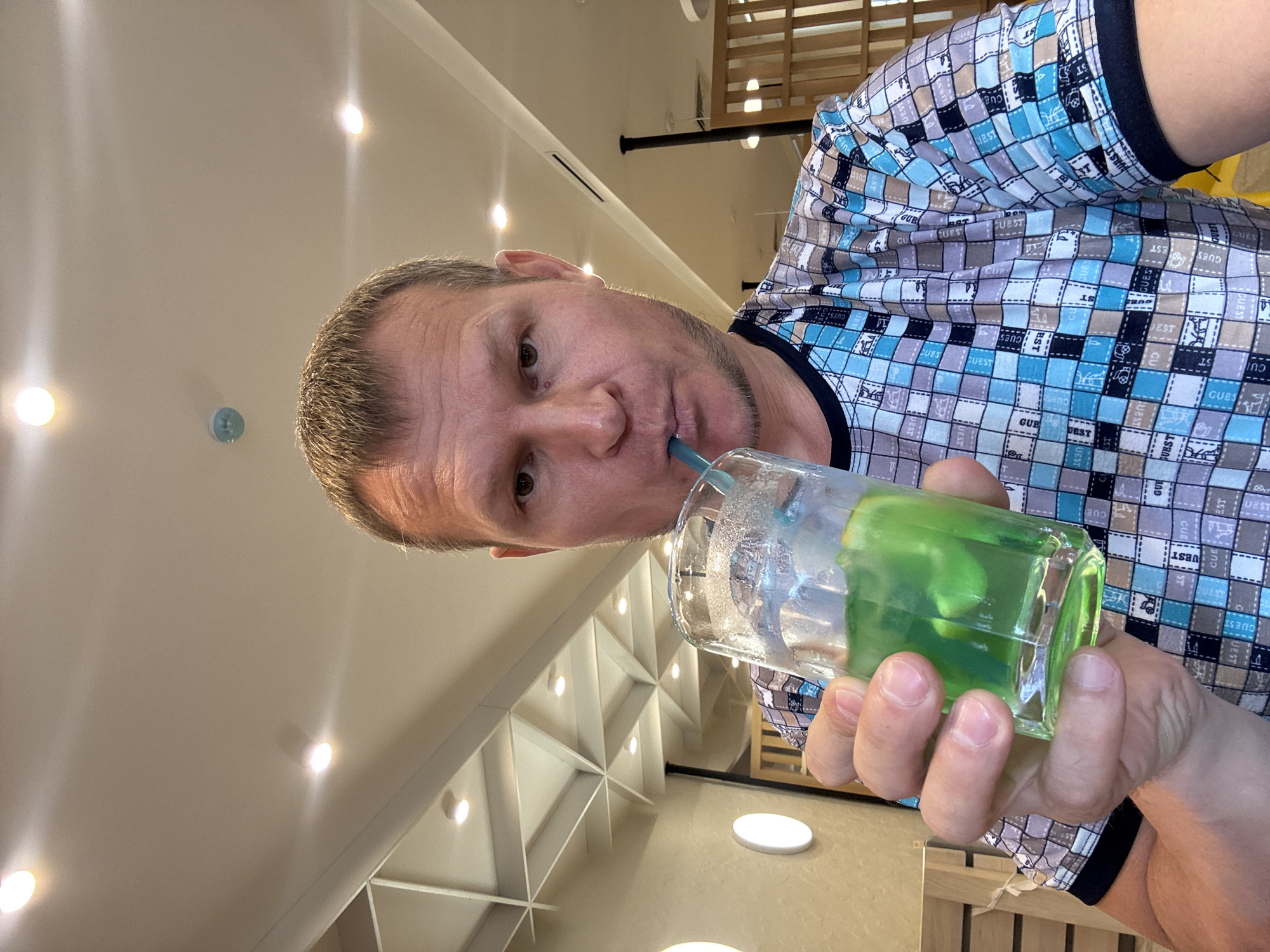 A man with short, blond hair is holding a glass of green liquid with ice and a straw to his mouth. He is wearing a t-shirt with a blue and brown checkered pattern. The background appears to be an indoor setting with a white ceiling featuring recessed lighting and a geometric design. The man's left arm is visible, holding the glass. The image is taken from a slightly low angle, looking up at the man. The location is identified as Oskemen, Kazakhstan.
