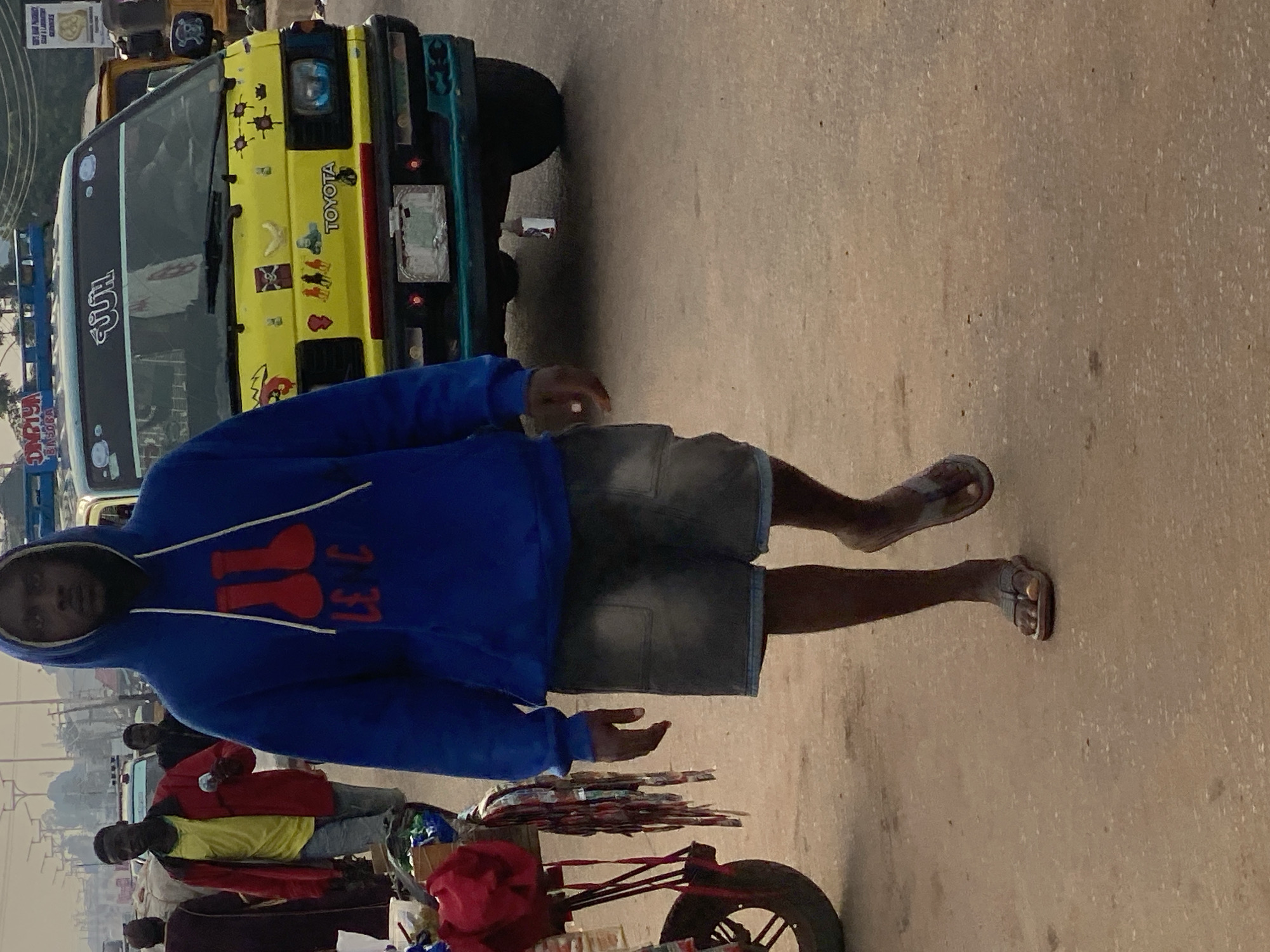 A low-angle, full-shot photograph captures a man walking on a dirt road in Sabon Tasha, Nigeria, during the day. The man, in the foreground, wears a blue hooded sweatshirt with a red graphic of two identical objects, resembling beakers, above text that appears to be "LE C". He also wears dark denim shorts and dark-colored flip-flops. To his left, a vendor's cart with a red cloth draped over it is partially visible, with various items, possibly goods for sale, hanging from its side. In the mid-ground, a yellow Toyota van, heavily decorated with stickers, including images of skulls and a banana, is parked. A man in a red jacket and yellow shirt stands near the van, holding a small object. Other individuals are visible in the background, along with other vehicles and structures that suggest an urban environment. The overall scene depicts a busy street market or roadside commerce area.