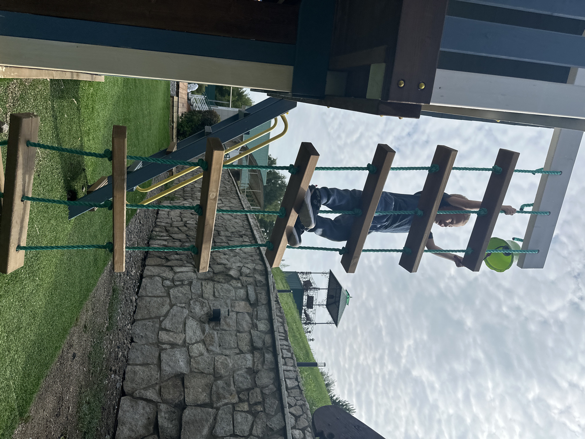 A young boy is climbing a rope ladder attached to a play structure. He is wearing a dark t-shirt and blue jeans. He is holding onto the rope with his right hand and appears to be reaching for the next rung. A green bucket is visible at the top of the ladder, held by the boy. The play structure is on a grassy area with a stone wall in the background. The sky is overcast with clouds. The location is indicated as Danilovka (Altai Krai), Russia.