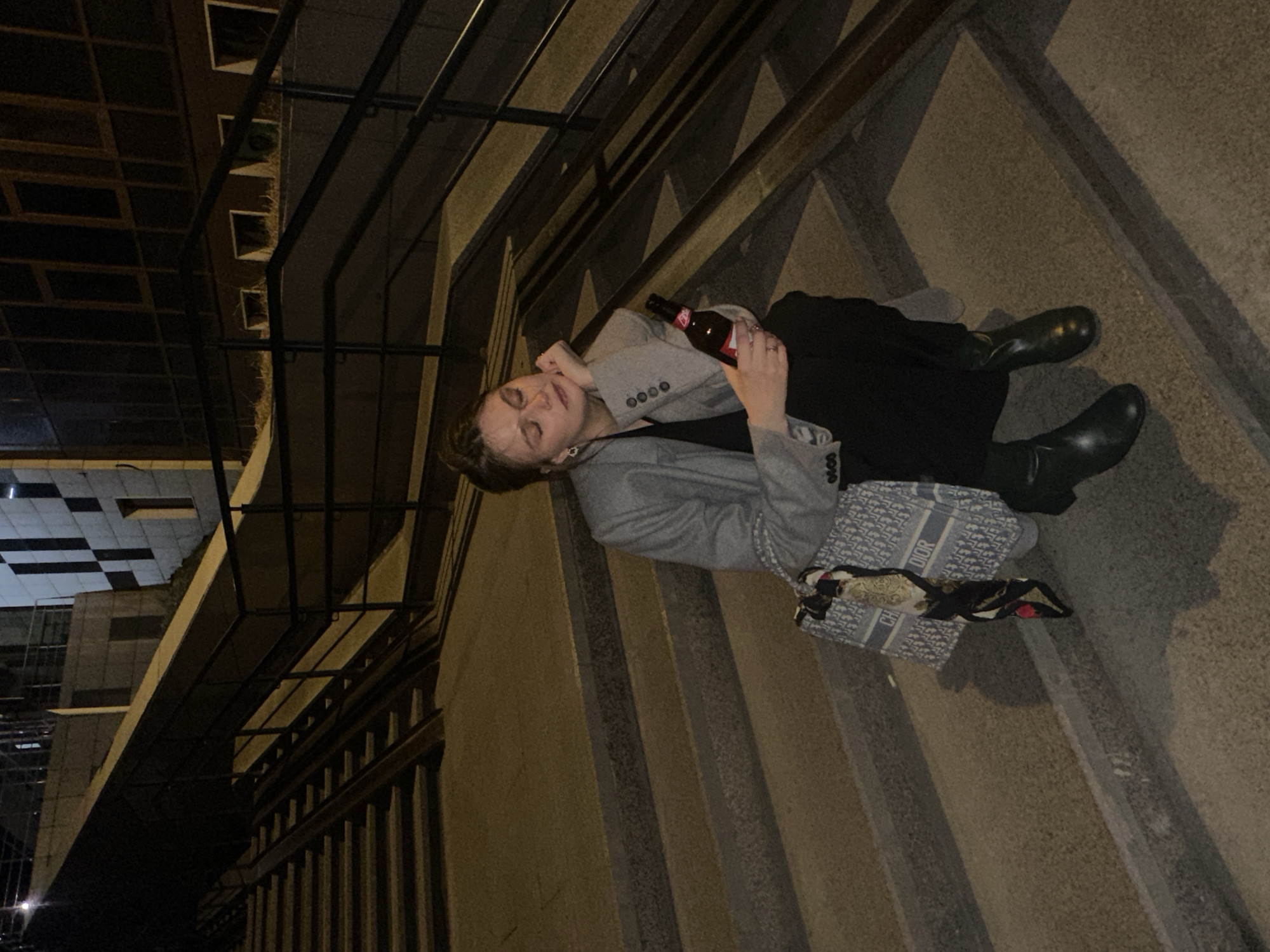 A young woman reclines on concrete steps outdoors at night. She wears black pants, black boots, and a grey blazer, and holds a red beer bottle in her hand. A blue and white patterned Dior bag lies beside her. The steps are part of an exterior staircase leading up to a building with a dark grid pattern on the upper floors. Iron railings run along the staircase. The woman is near a building. The building's location in the city of Krasnoyarsk, Russia is not discernible from the image.