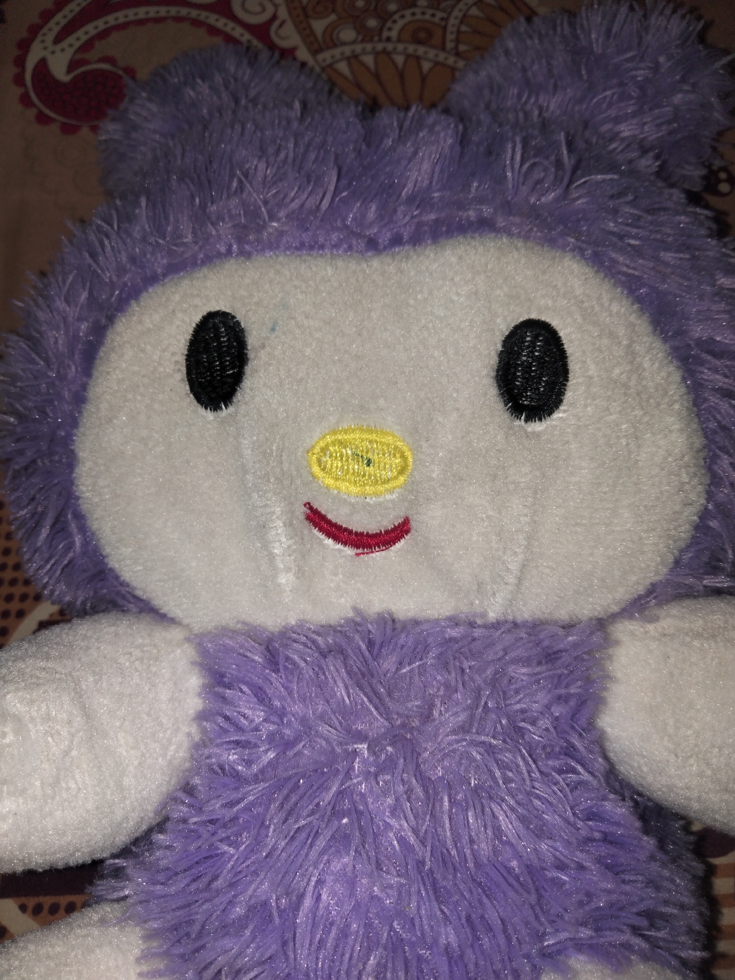 A close-up, head-on, full-body shot shows a stuffed toy. The toy is primarily purple and white, with black eyes, a yellow nose, and a red mouth. The background is a patterned fabric, possibly a bedsheet or blanket. The image appears to have been taken in an indoor setting, with no discernible elements indicating its location within the city of Payagaboh, Indonesia.