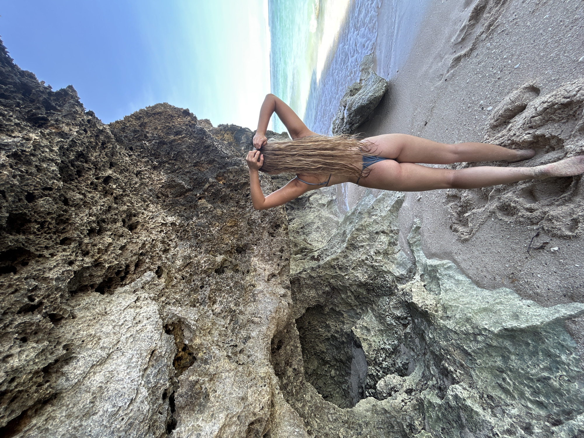 A woman in a bikini stands on a beach near a rocky outcrop in Kuta Selatan, Indonesia. She has long, blonde hair that reaches her lower back and is arranging her hair with both hands. She is positioned between a rough, porous rock formation on the left and the beach on the right. The sand on the beach is light tan. The ocean is visible in the background, meeting the horizon under a clear, blue sky.