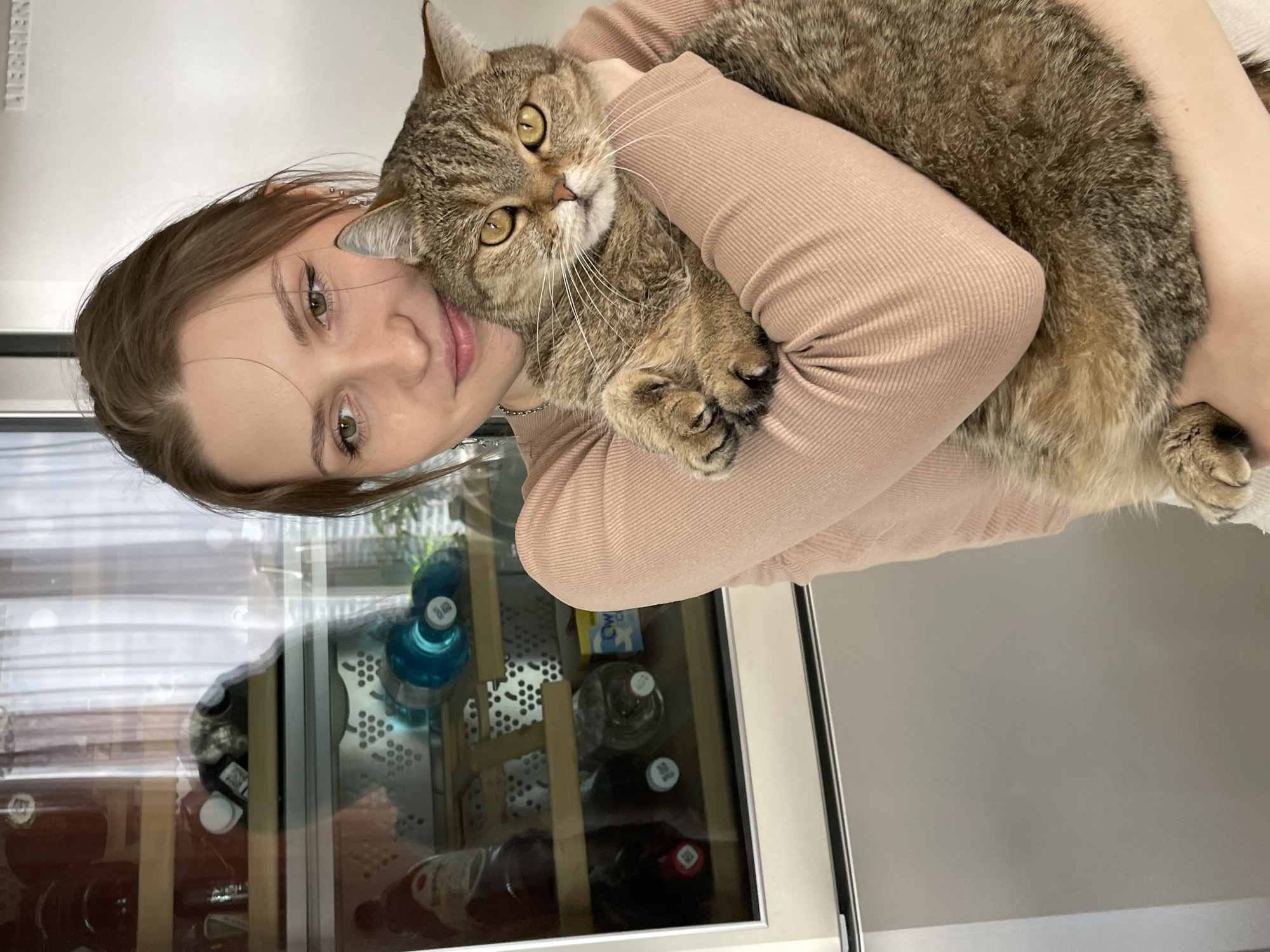 A young woman is captured in a close-up portrait, holding a tabby cat in an indoor setting. The image is oriented vertically, showing the woman's head and upper torso, with the cat resting comfortably against her chest and shoulder, looking directly at the viewer.
The woman has fair skin, long brown hair that cascades around her shoulders, and striking green or hazel eyes. Her lips are a soft pink, and she wears a subtle, gentle smile, conveying warmth and affection. She is dressed in a light-colored, possibly beige or light pink, long-sleeved top with a ribbed texture.
The cat is a domestic short-haired tabby with prominent brown and black stripes and light-colored paws. Its large, luminous golden-yellow eyes are wide open, fixed on the camera, and it has long, delicate white whiskers. The cat appears relaxed and well-cared for, nestled securely in the woman's arms.
In the background, a modern wine cooler or refrigerator with a glass door is visible. Through the glass, various bottles can be seen, including dark-colored wine bottles and clear plastic water bottles, some with blue caps. A distinctive white patterned surface and wooden dividers are also visible inside the cooler. Above the cooler, a light gray or white wall extends. Text that reads "LIEBHERR" is discernible on a metallic emblem or label on the wall or appliance.
The scene exudes a sense of comfort and a loving bond between the woman and her pet. The lighting suggests an indoor setting, possibly during the daytime. This intimate moment is described with the provided context of Saint Petersburg, Russia.