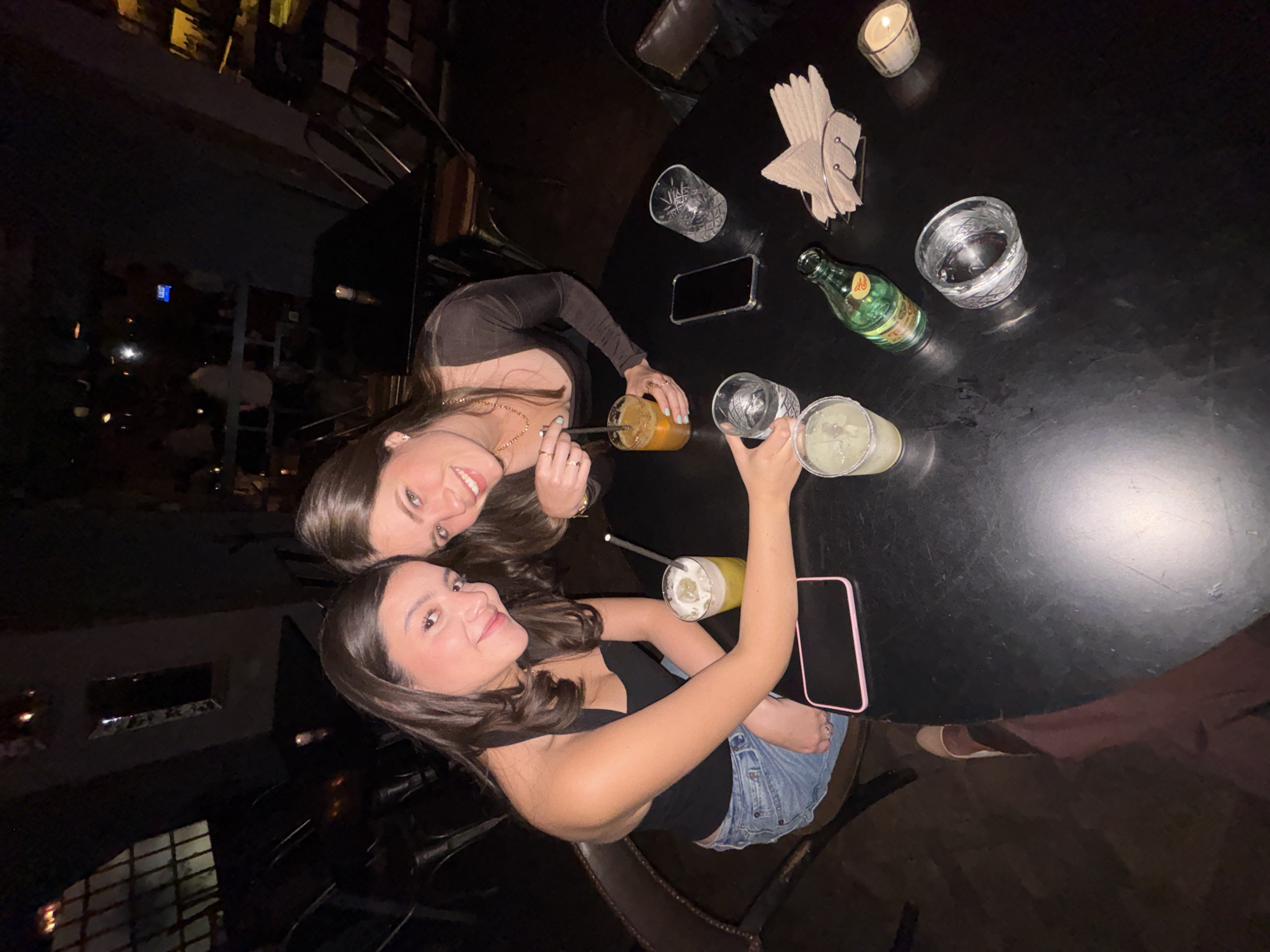 Two women are pictured at a table at a bar, location Unknown, Unknown. The woman on the left is wearing a black long-sleeved shirt and a gold necklace and drinking an orange drink. The woman on the right is wearing a black tank top and jean shorts and holding a clear glass. There are drinks, a phone, a bottle of Topo Chico, napkins, and a candle on the table.