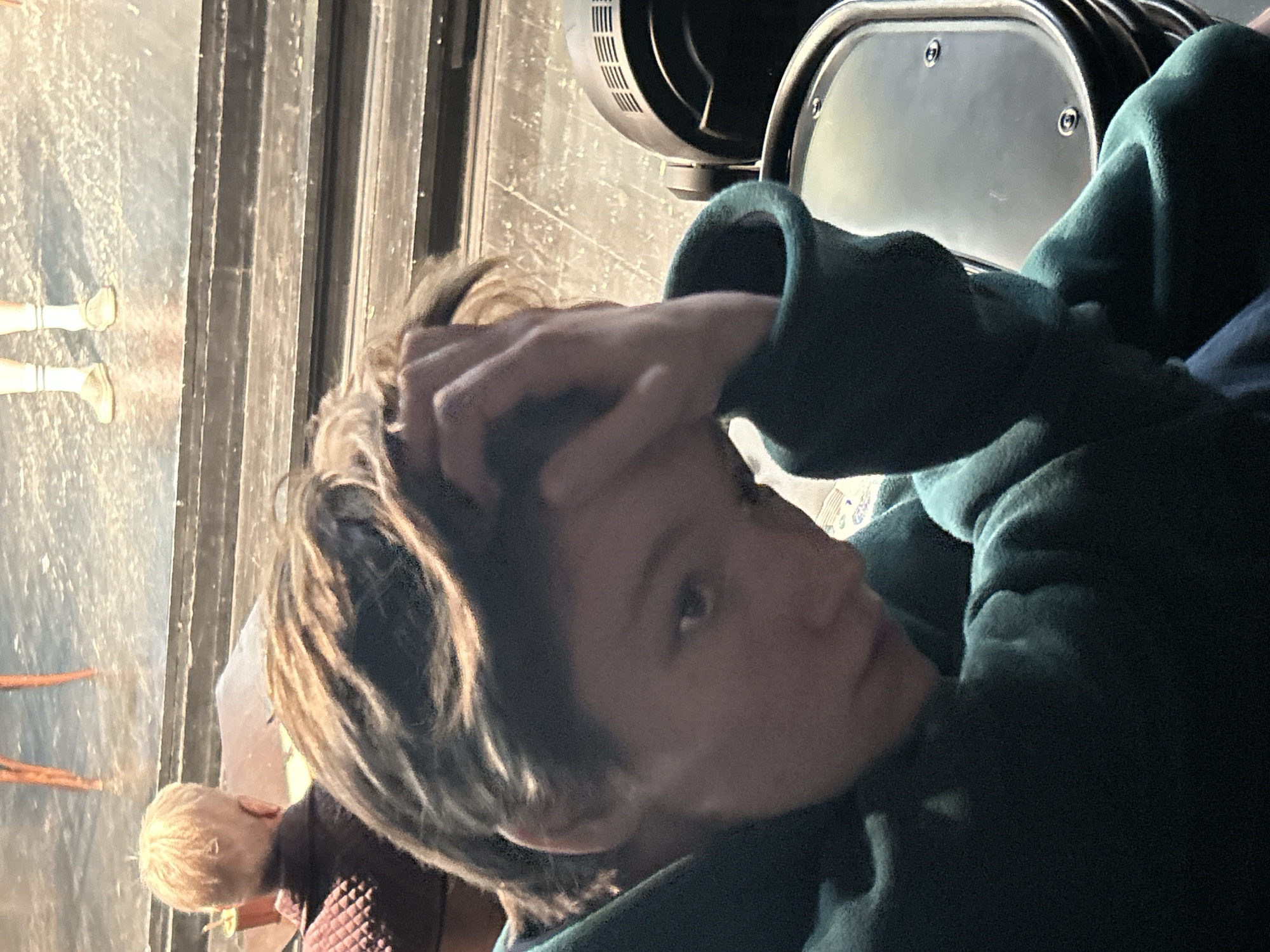 The image, rotated counter-clockwise, depicts an indoor scene with several individuals and equipment. In the foreground, a young person with light brown and blonde hair, wearing a dark green hooded sweatshirt, is positioned prominently. They are looking towards the left side of the frame, with their left hand raised and resting on their forehead or in their hair.
Behind the main subject, to the left, the back of a small child's head with blonde hair is visible, wearing a dark top. Further into the background, also on the left, two pairs of legs clad in light-colored footwear (possibly boots or shoes) are standing on a light-colored, textured surface that resembles a stage floor. These legs appear to be part of a mannequin or someone standing very still.
On the right side of the background, several dark, metallic objects are partially visible. One object has vents, suggesting it might be a fan or part of a machinery unit, while another is a rounded metallic surface with visible bolts, possibly part of equipment or a seat. The background also shows vertical dark lines, which could be stage curtains or panels. The lighting is dim but highlights the main subject's hair. No text is visible in the image.