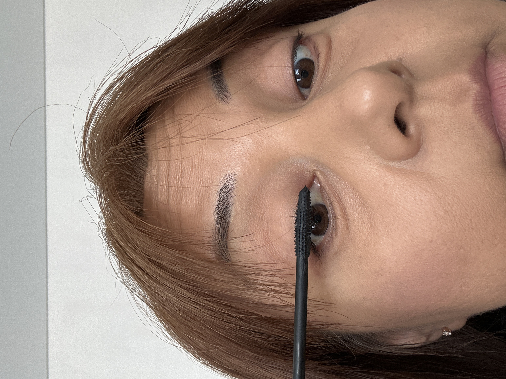 A close-up, indoor photograph depicts a woman applying mascara to her lower eyelashes with a black wand applicator. The woman has brown hair and brown eyes, and her skin appears fair. The background is a plain, light-colored surface, possibly a wall. The location is unknown.