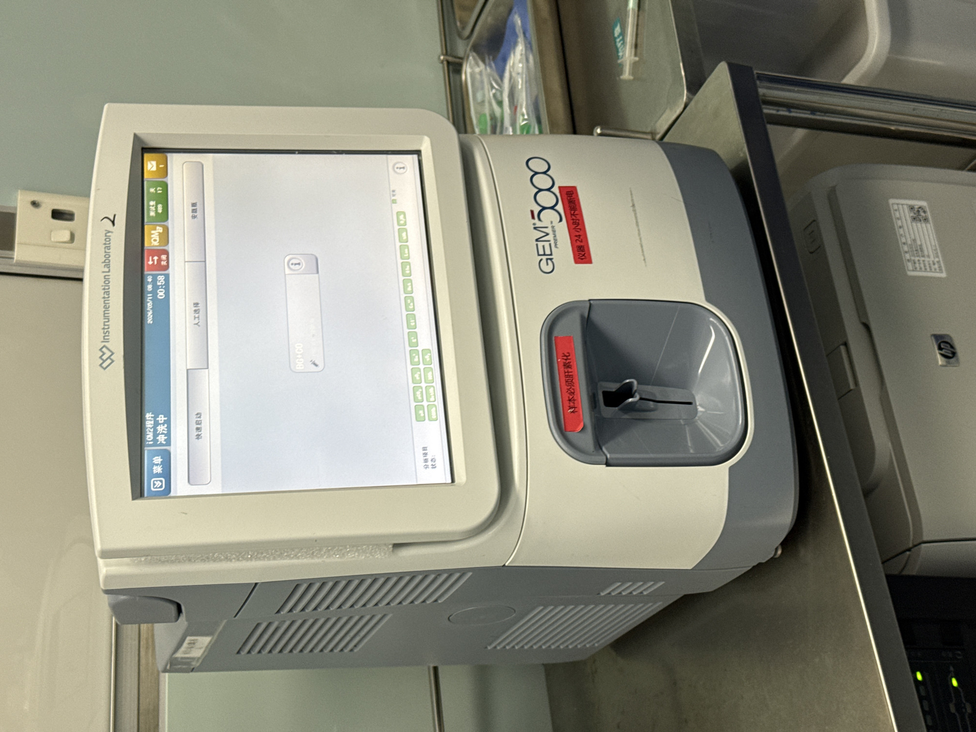 The image shows a GEM Premier 5000 blood gas analyzer. Its screen displays Chinese text and an analysis menu. The analyzer is located on a stainless steel counter next to a white printer. The image lacks sufficient context to place the scene in the city of Yuzhong District, China.