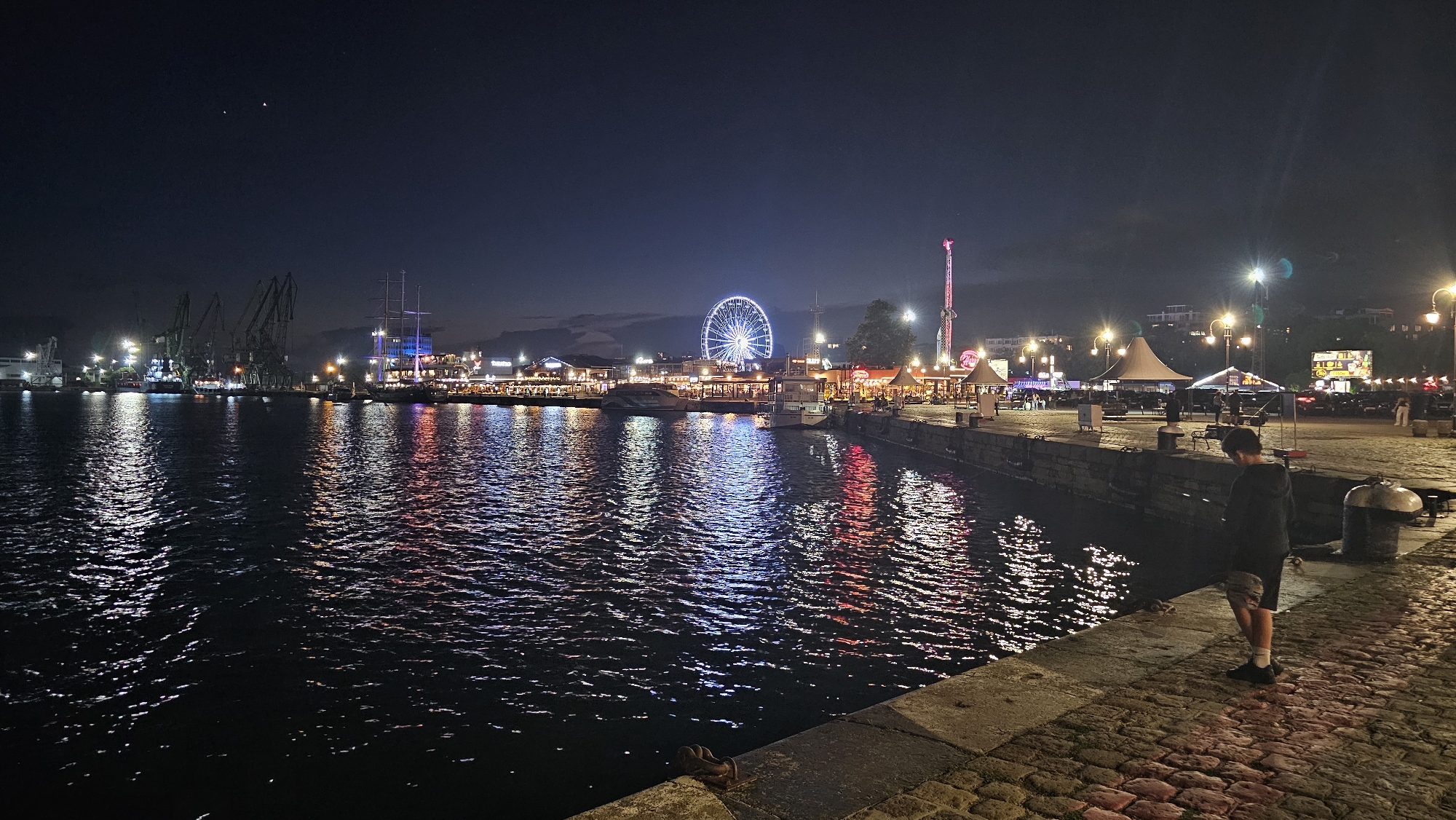 The image depicts a night scene at the port of Varna, Bulgaria, specifically along the waterfront promenade. In the foreground, a person stands facing away from the camera, looking towards the water. The promenade is paved with cobblestones and illuminated by streetlights. The water in the port reflects the vibrant lights of the city, including those from a Ferris wheel and amusement park attractions visible in the middle ground. Further back, industrial cranes stand silhouetted against the dark sky, indicating the active port environment. Buildings with illuminated windows line the waterfront, and various boats are docked. The sky is dark, suggesting it is nighttime, with a few faint stars visible.