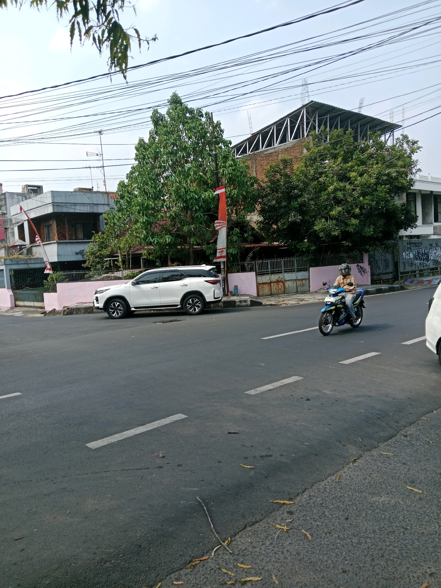 The image depicts a street scene in Ciamis, Indonesia. A white SUV is parked on the side of the road. In the foreground, a person wearing a brown shirt and helmet is riding a blue and yellow motorcycle. The street is paved and has white lane markings. Buildings, trees, and power lines are visible in the background. The sky is clear and bright.