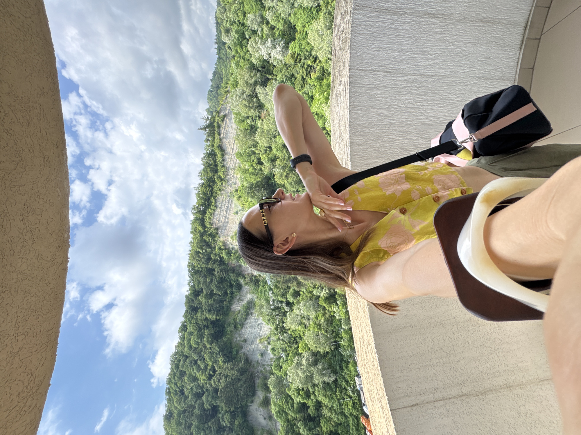 This vertical, eye-level media file shows a woman relaxing on a balcony overlooking a lush, green mountainside. She is leaning back, with her right arm extended upwards and her hand near her face, and her left arm resting on her lap. She is wearing a yellow floral-patterned dress with buttons down the front and a black watch on her left wrist. She also has on sunglasses with the word "GUCCI" visible on the side. A black bag with pink accents is positioned behind her on the balcony. The balcony itself has a textured beige railing and a lighter-colored wall behind her. The background is dominated by a steep, verdant mountain covered in trees, with visible rocky outcrops. The sky above is blue with scattered white clouds, suggesting it's a bright, clear day. The woman's pose and facial expression suggest relaxation and enjoyment of the scenic view. The location context is Sochi, Russia.
