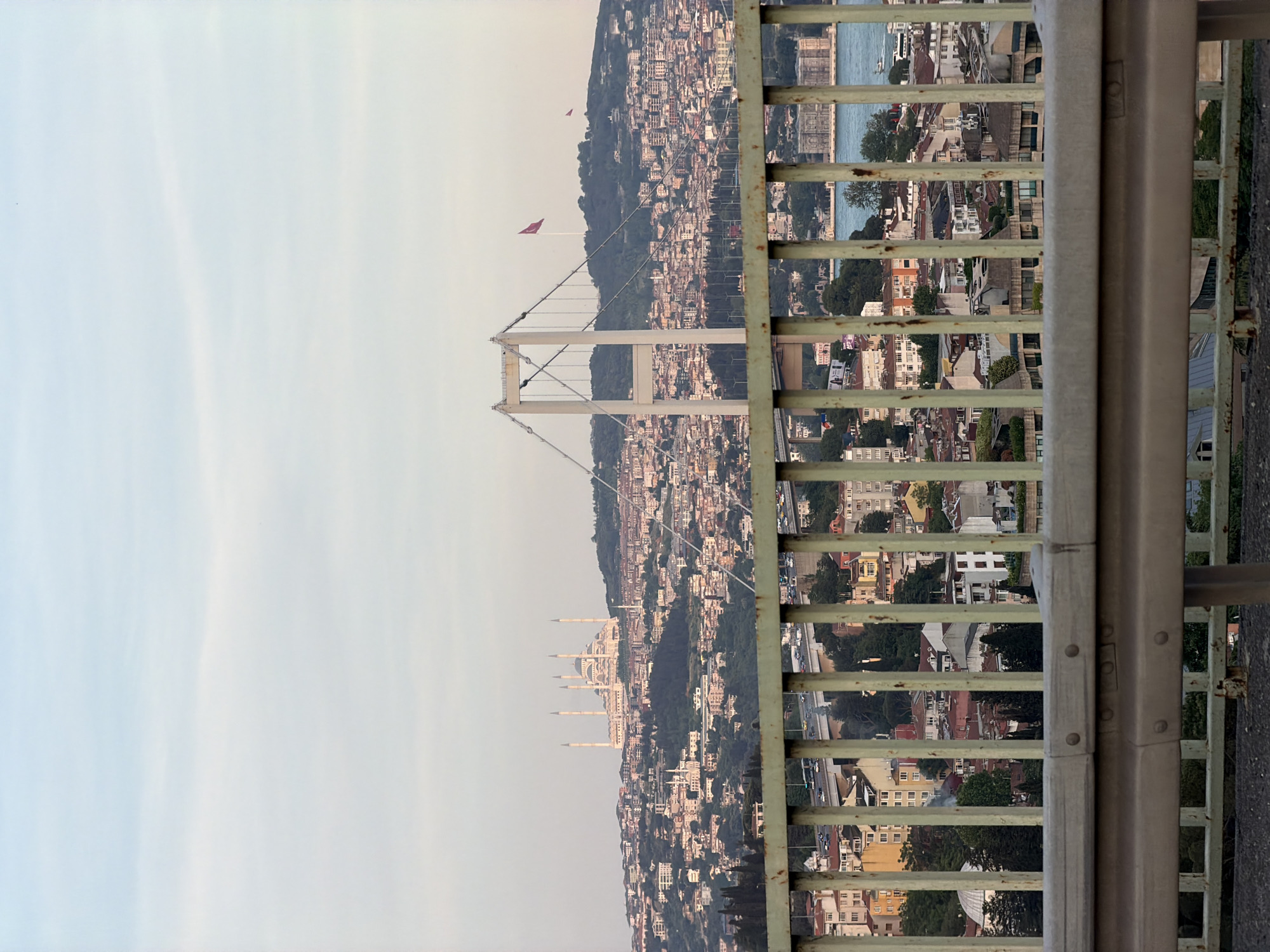 The image displays a panoramic daytime view of a sprawling city built across hillsides, overlooking a wide body of water, likely the Bosphorus Strait in Istanbul. The perspective is elevated, with a metal guardrail or fence prominently occupying the immediate foreground, partially obscuring the lower cityscape.
Beyond the railing, a dense urban environment unfolds, characterized by countless buildings, mostly residential, covering green, undulating hills. In the midground, a large suspension bridge, featuring two distinct white towers, spans across the water. Further to the left, a grand mosque with multiple tall, slender minarets stands majestically on a hilltop, serving as a prominent landmark against the horizon. Two red Turkish flags are also visible on flagpoles on distant hills.
Through the gaps in the foreground railing, glimpses of the city below are visible, including more buildings, roads with moving vehicles, and boats traversing the water. The sky is bright but appears somewhat hazy or softly overcast, suggesting a time of day like late afternoon or early evening. The scene conveys the vastness and architectural density of a major metropolis. There is no specific human activity highlighted, nor is any legible text visible on structures or flags. The overall impression is one of a grand urban landscape.