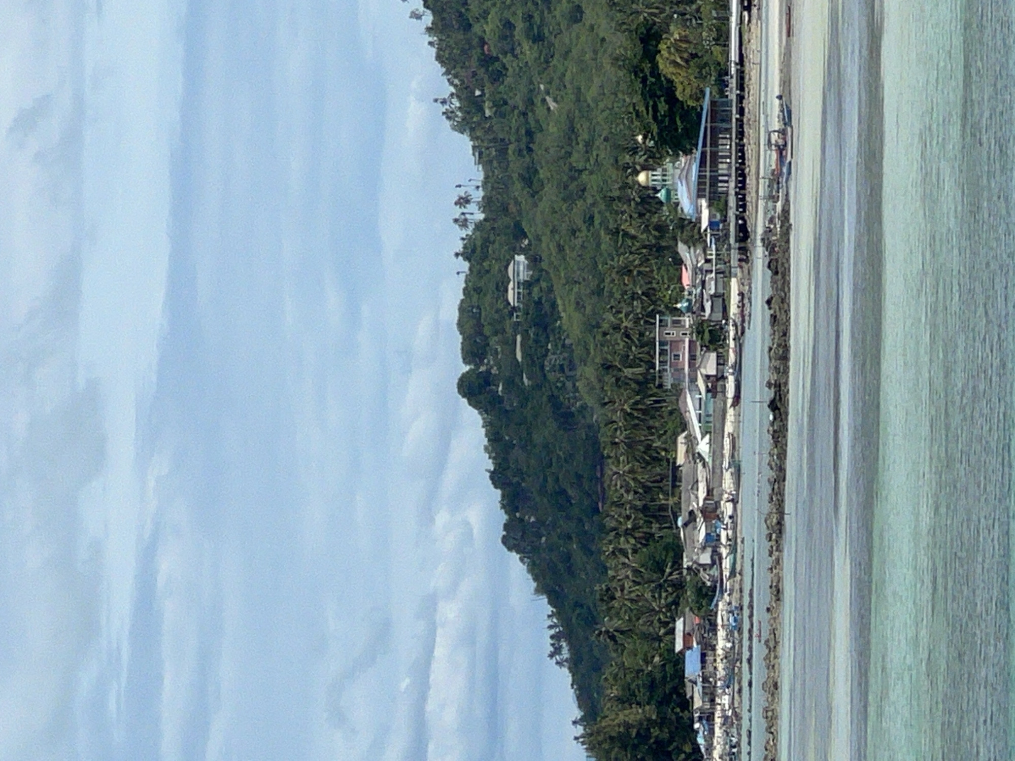 The image shows a coastal village on the island of Ko Samui in Thailand. In the foreground, the turquoise waters of the sea are visible, with a breakwater made of rocks stretching across the water. A few small boats are anchored near the shore.
In the middle ground, a collection of buildings forms the village. These include houses, shops, and what appears to be a mosque with a prominent golden dome. The buildings are situated along the coastline, with a road or pathway running behind them. Palm trees and other lush vegetation are abundant, extending up the hillside behind the village.
The background is dominated by a lush green, forested hillside that rises steeply. Several houses are visible nestled amongst the trees on the slope. The sky is partly cloudy, with patches of blue sky showing through. The overall scene depicts a tropical coastal settlement with a blend of natural beauty and human habitation.