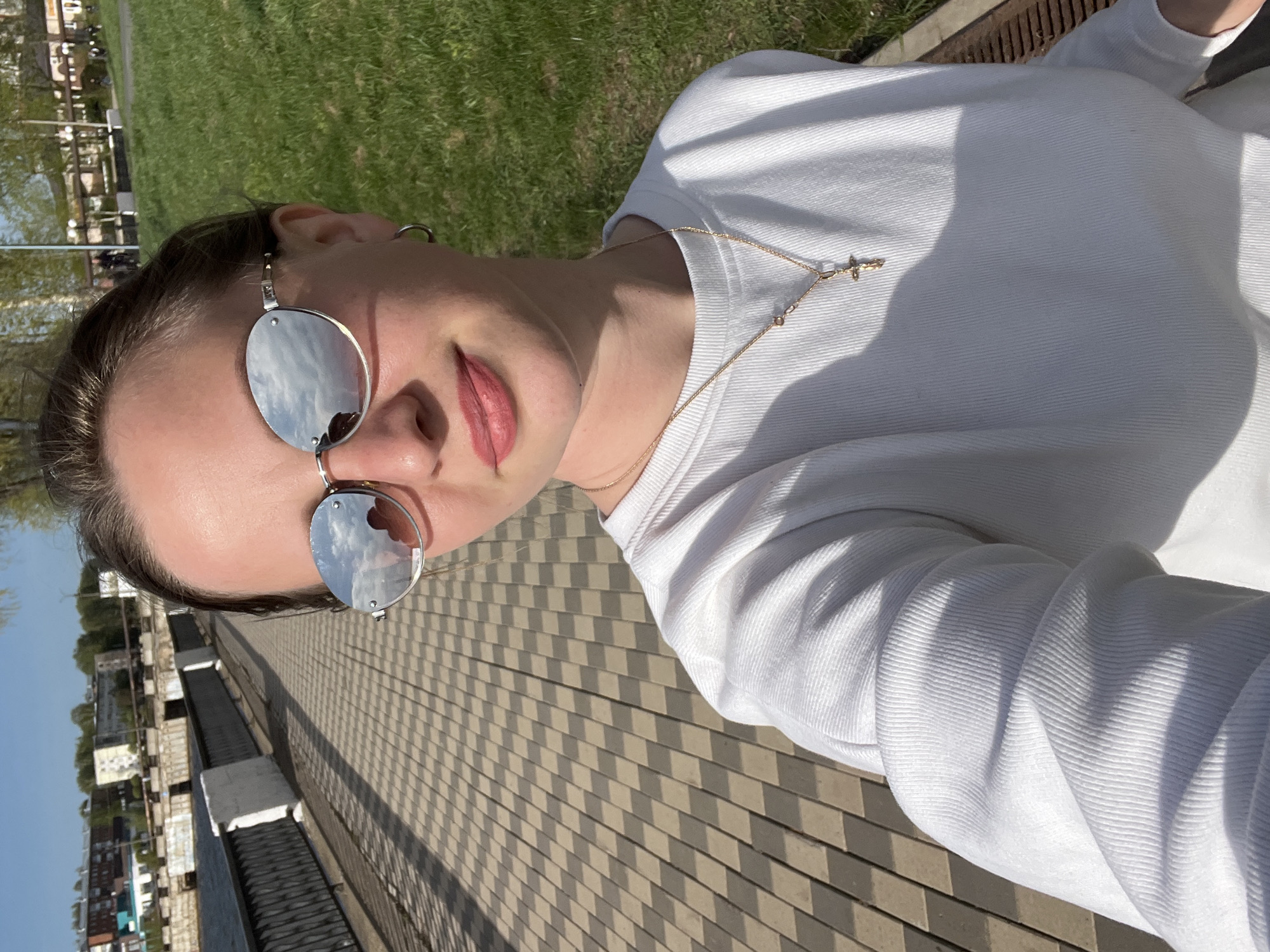 A selfie of a young woman outdoors on a sunny day. She is wearing a white long-sleeved shirt, a gold necklace with a cross pendant, and round sunglasses that reflect the sky and clouds. She is standing on a light-colored, patterned surface. In the background is a body of water with buildings and trees along the shore. Based on the architecture visible in the background, this location is likely the city of Votkinsk, Russia. There is also a patch of green grass and a metal grate visible.