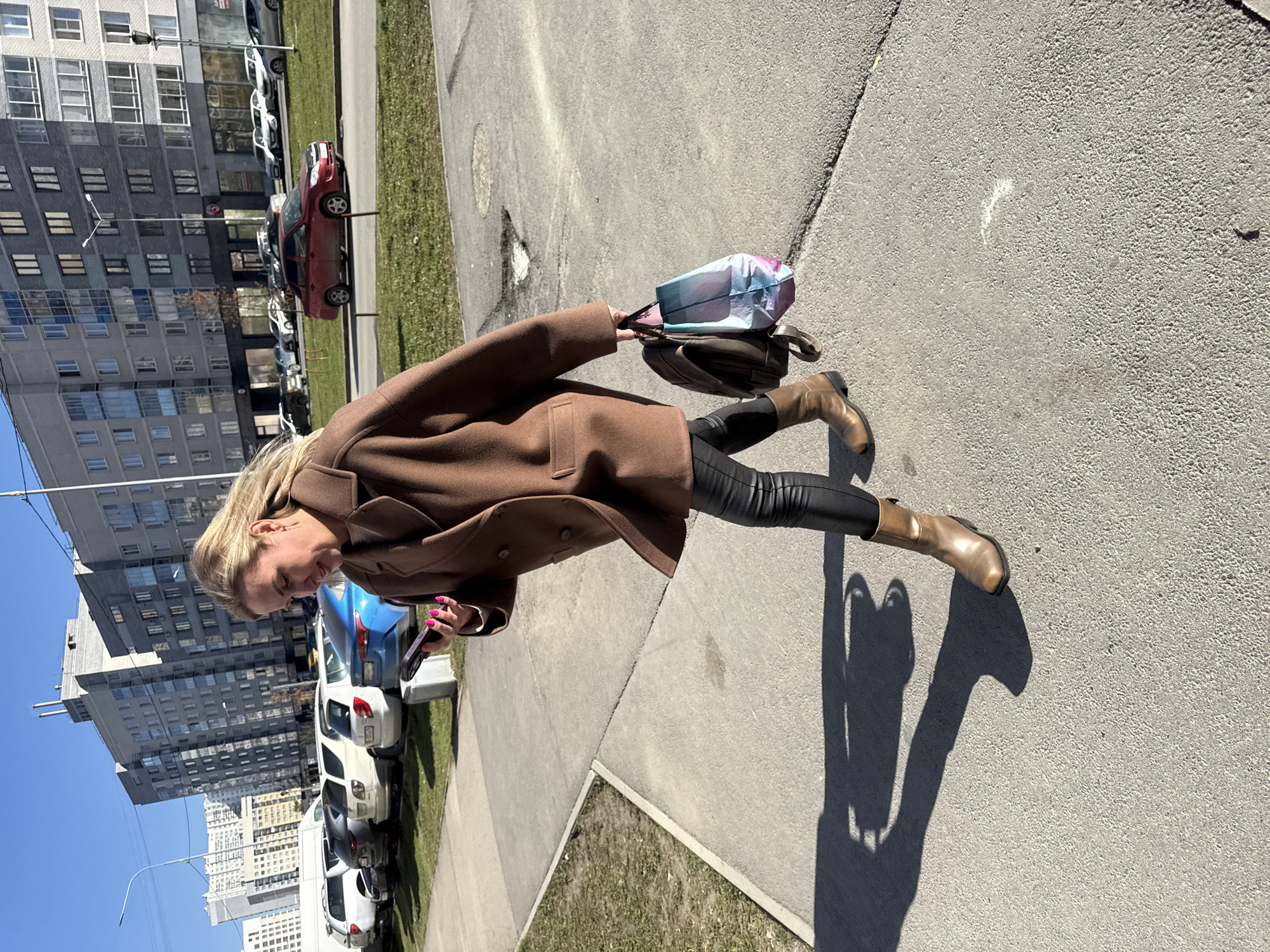 The image shows a woman walking on a sidewalk in Yekaterinburg, Russia. She is wearing a brown coat, black pants, and brown boots. She is holding a phone in one hand and a bag in the other. The background includes buildings, parked cars, and street vegetation. The image is taken during daylight.