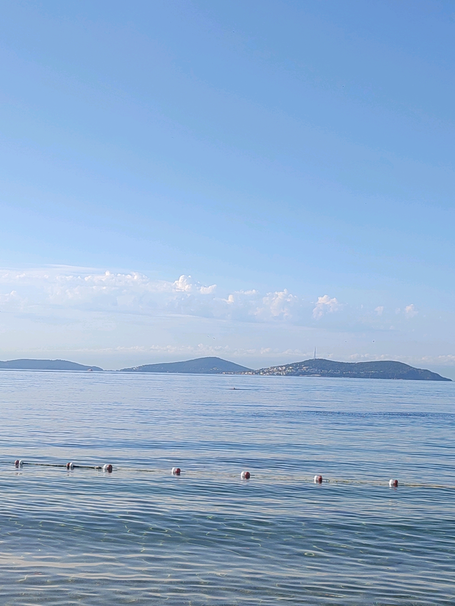 The image depicts a calm, serene body of water, likely the Sea of Marmara, seen from a beach in Kadıköy, Turkey. In the foreground, a rope with buoys floats on the surface, marking a boundary in the clear, shallow water where the sandy seabed is visible. The water exhibits gentle ripples and reflects the soft blue of the sky. In the mid-ground, a calm expanse of sea stretches towards distant islands. One island, with a settlement and a prominent tower, is clearly visible, while other islands appear as darker silhouettes against the horizon. The sky above is a pale, cloudless blue, transitioning to lighter shades near the horizon. The overall scene conveys a sense of tranquility and natural beauty characteristic of the coastal areas of Kadıköy.