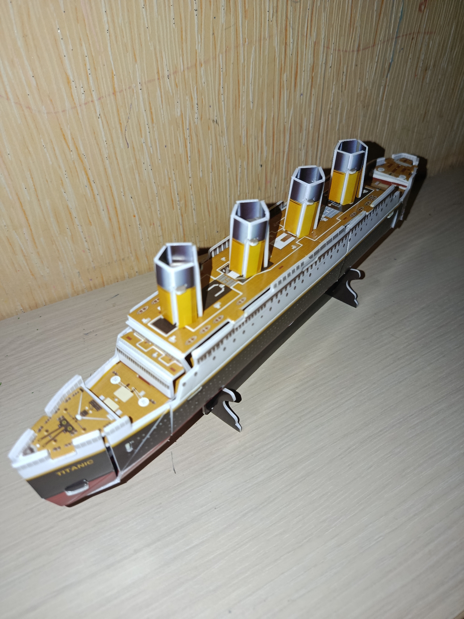 A 3D paper model of the RMS Titanic is displayed on a light-colored, wooden surface. The model is positioned at an angle, with its bow pointing towards the lower left of the frame. The ship features its characteristic four funnels, painted yellow with silver tops, and a white hull with a dark grey bottom. The deck is detailed with various structures and markings. The background consists of a textured wooden wall, with vertical grain patterns and subtle variations in color. There are no people present in the image, and the scene appears to be indoors, on a shelf or table. The lighting suggests an indoor setting, possibly with artificial light. There is no identifiable information to suggest a specific location in Chișinău, Moldova.