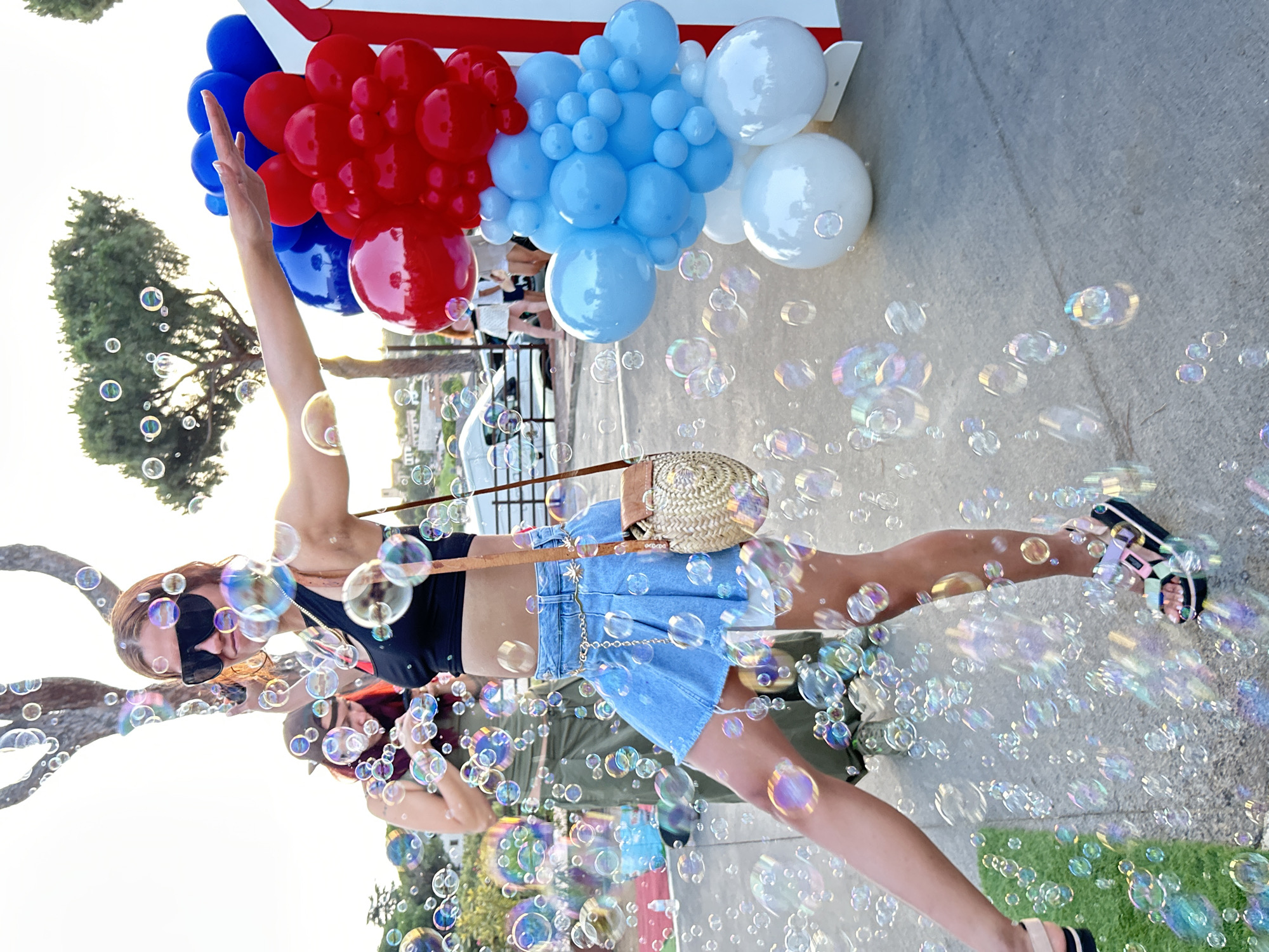 The image depicts a woman in a black crop top and a blue denim skirt, adorned with a gold chain belt, standing amidst a flurry of soap bubbles. She is wearing oversized black sunglasses and multi-colored sandals. Her left arm is extended, and her right arm is bent, holding a straw purse. She is positioned in the foreground, with another woman in a green outfit visible behind her. The background features trees, a street with parked cars, and a balloon arch composed of red, blue, and white balloons. The scene appears to be an outdoor event, possibly a celebration or party, in the city of Sant Vicenç De Montalt, Spain, indicated by the context of the request.