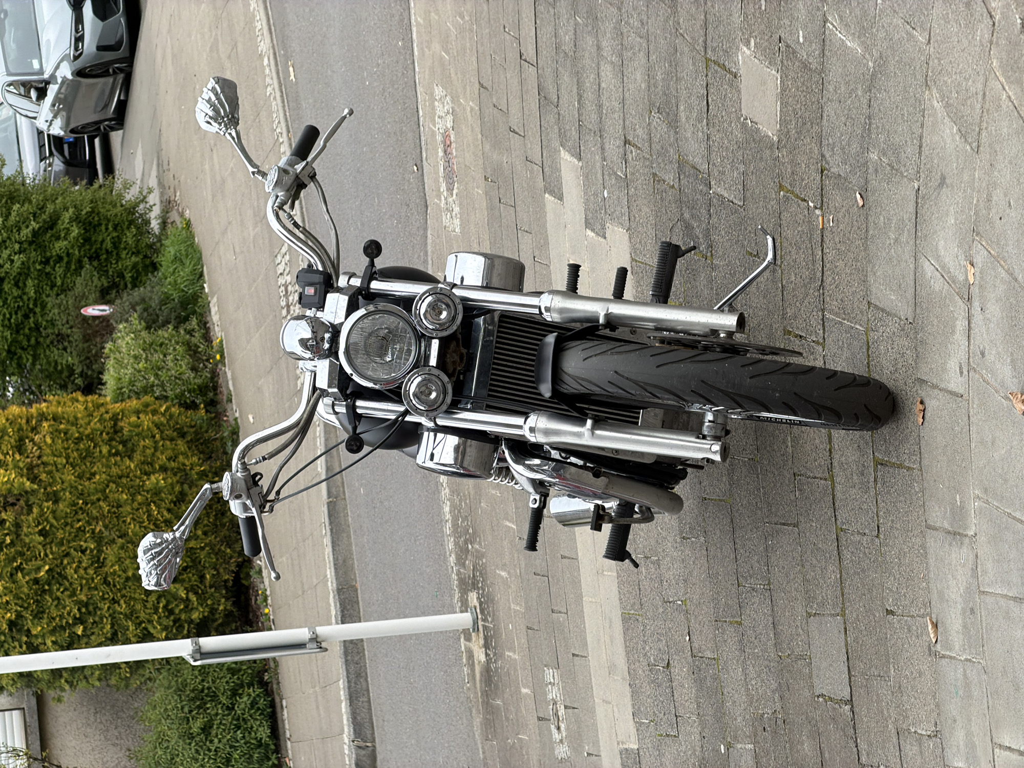 The image shows a motorcycle parked on a paved surface, possibly a sidewalk or parking area, in an urban environment. The motorcycle is facing the viewer, providing a clear view of its front end. The handlebars are raised, with chrome skeleton-hand-shaped grips. The motorcycle's headlights are prominent. A car is visible in the background, and greenery suggests a residential area. There are no people visible in the immediate vicinity of the motorcycle. The image was taken in Esch-sur-Alzette, Luxembourg.