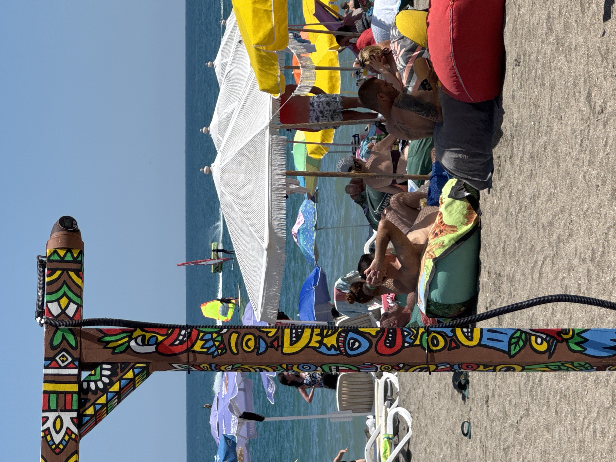 The image depicts a sunny day at the beach in Burgas, Bulgaria. In the foreground, a wooden pole adorned with colorful, tribal-like paintings stands vertically. To the right of the pole, several people are relaxing on beanbag chairs on the sandy beach. The sea is visible in the background, with clear blue water under a bright sky. Windsurfers are seen on the water, indicating recreational activity. Various beach umbrellas, mostly white and yellow, are spread out, providing shade for beachgoers. Some individuals are seated or reclining under the umbrellas, enjoying the beach environment. The overall scene suggests a lively and relaxed atmosphere at a popular beach destination.