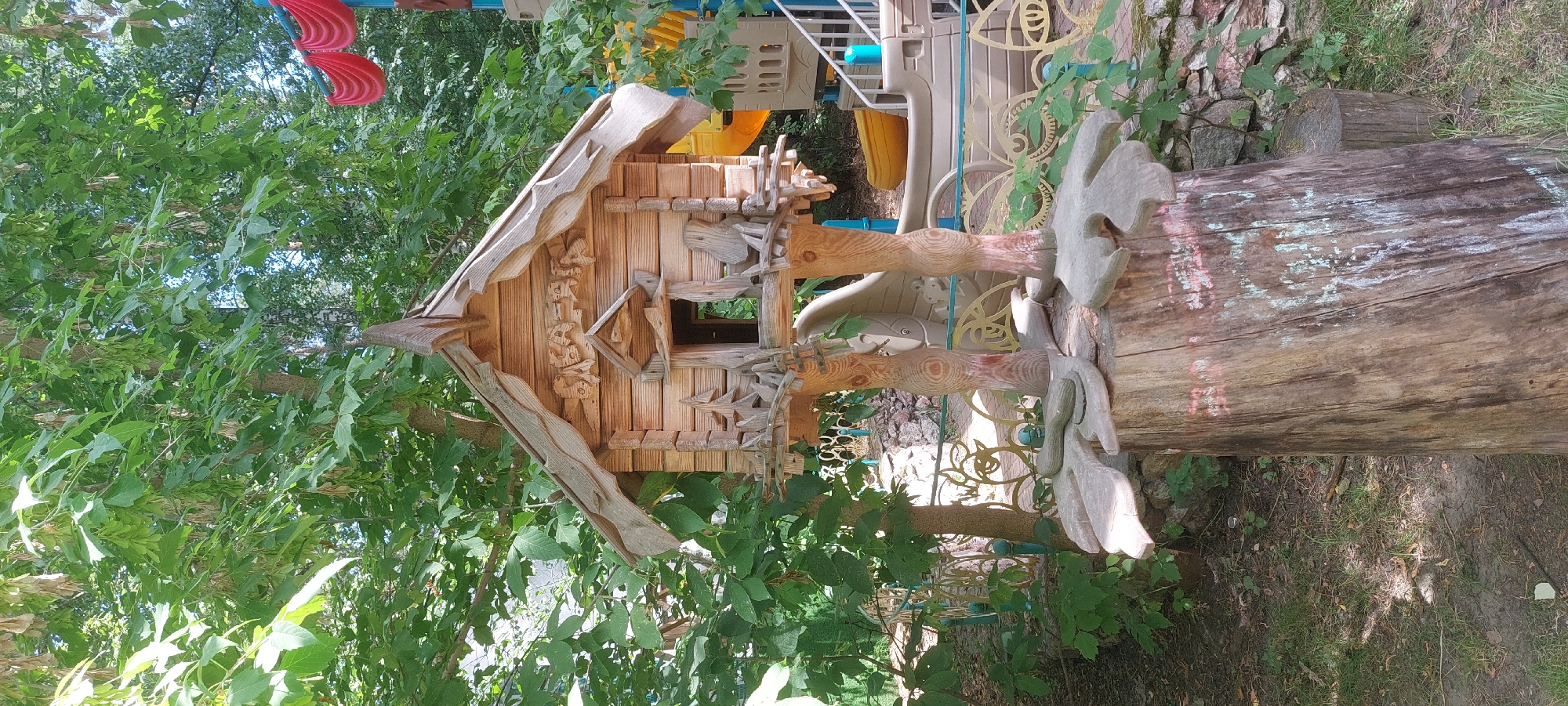 A whimsical, hand-carved wooden birdhouse, designed to resemble a miniature fairytale dwelling, stands as the central subject of this outdoor scene. The birdhouse is elevated on two intricately carved wooden legs that are anchored to a large tree stump. The stump itself is adorned with faded chalk drawings, suggesting it may have served as a canvas for children's art. The birdhouse features a detailed, gabled roof with decorative carvings, a small open doorway, and tiny wooden fences. Surrounding the birdhouse, lush green foliage from trees and bushes creates a natural, verdant backdrop. In the background, elements of a playground are visible, including colorful structures, slides, and railings, indicating the setting is likely a park or recreational area within the city of Berdsk municipality, Russia. The lighting suggests it is daytime, with sunlight filtering through the leaves. There are no people present in the image.