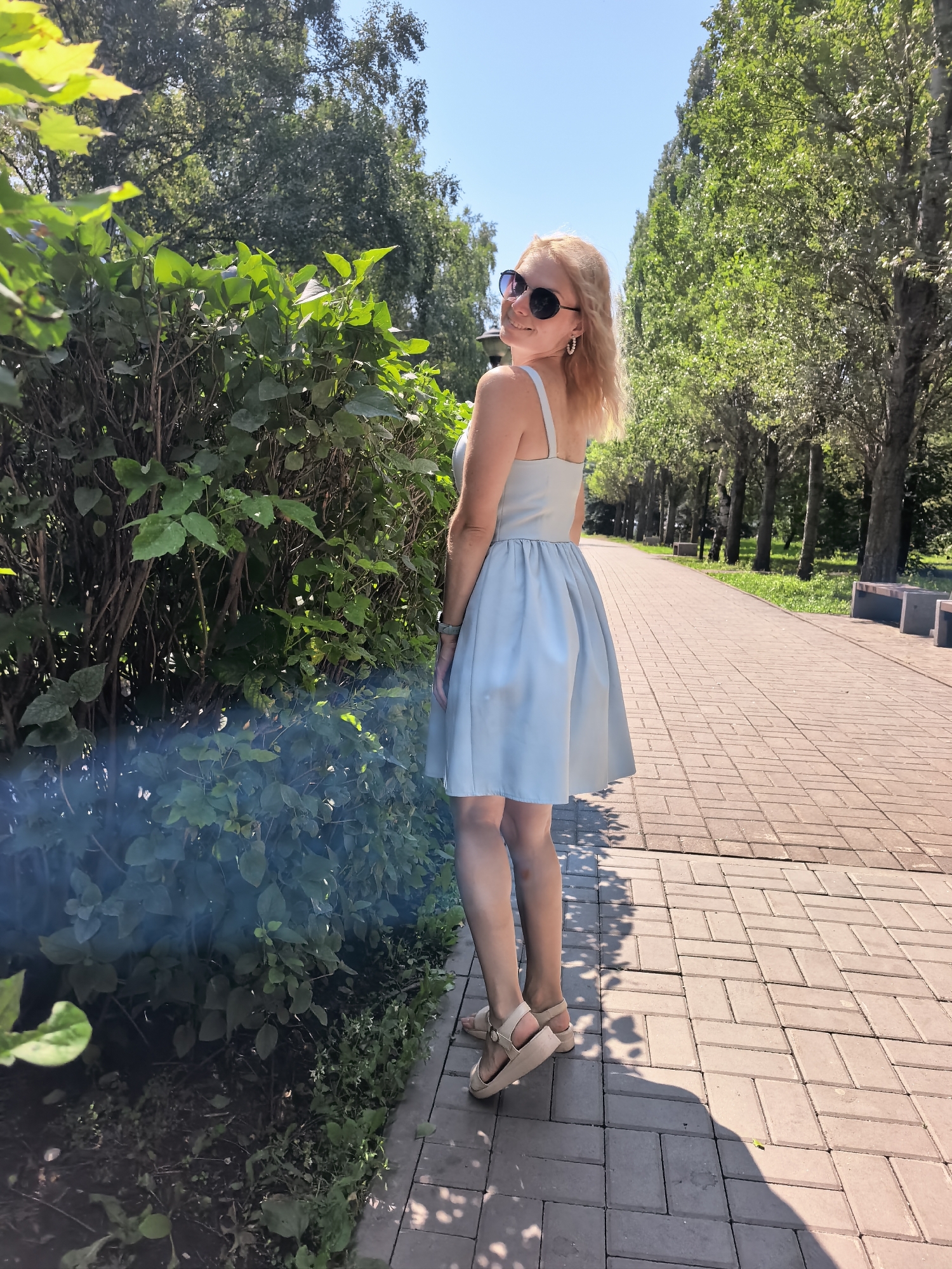 A woman with blonde hair, wearing sunglasses and a light blue dress, poses in a park with a paved walkway and trees. She is standing on the walkway, which is made of rectangular pavers. To her left is a dense bush, and to her right, the walkway extends into the distance, lined with trees and benches. The sky is clear and blue. The scene appears to be in Sterlitamak, Russia, based on the provided context, likely a park or public garden area.