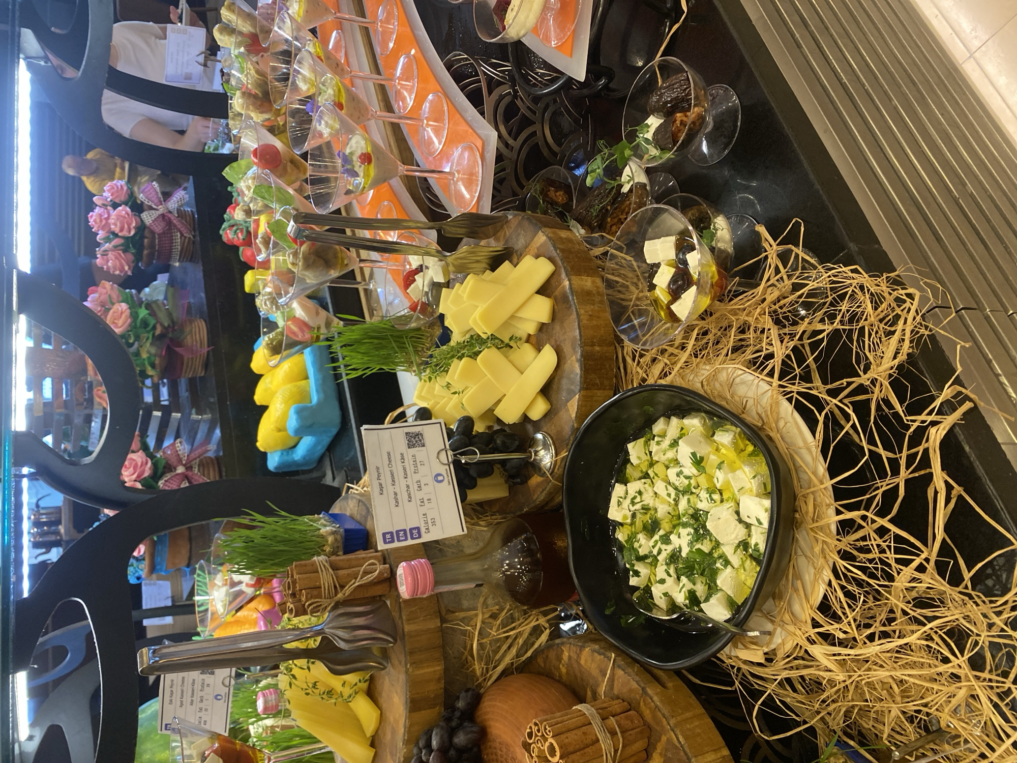 The image depicts a buffet table, possibly in Aksu, Turkey, featuring a variety of food items. In the foreground, a black bowl contains diced white cheese with herbs and olive oil, placed on a bed of straw. To the left of the bowl is a round, dark object, possibly cheese or bread, with cinnamon sticks bundled next to it. Various cheeses are displayed on wooden platforms, including sliced yellow cheese and a round of cheese. A bottle with a pink cap and dark liquid is positioned between the cheese displays. In the mid-ground, small appetizer portions are served in martini glasses, containing what appears to be dips, olives, and other garnishes. Green sprouts in small blue containers are also visible. In the background, a buffet setup with decorative floral arrangements is present, and two individuals are partially visible, one with their back to the camera and another seated. The lighting suggests an indoor setting, possibly a restaurant or hotel.
