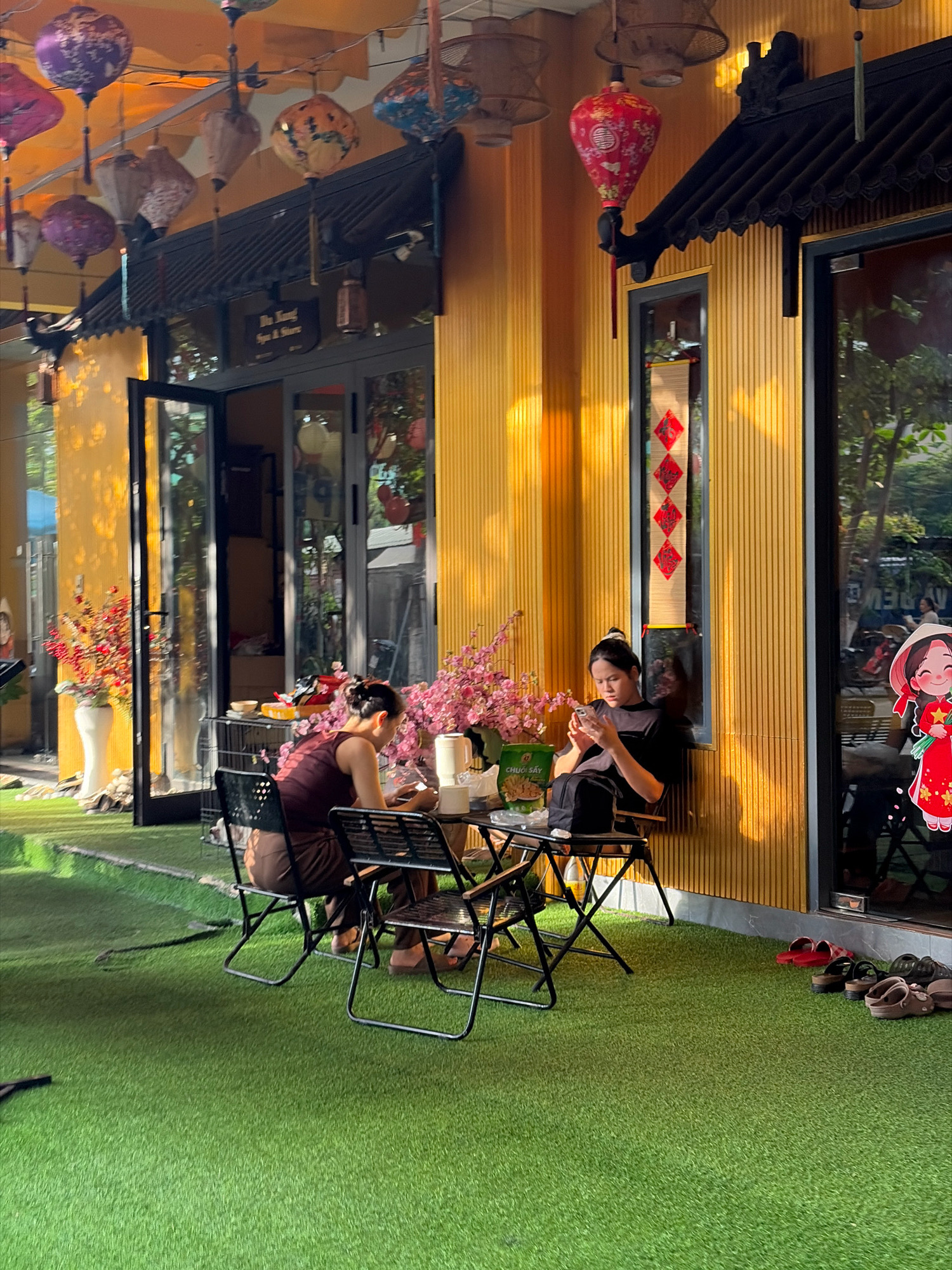 The image depicts an outdoor seating area of a cafe or shop located in Đà Nẵng, Vietnam, likely on a street or pedestrian walkway given the vibrant, decorated facade. The building exterior is painted a bright yellow with vertical ribbed siding and features large glass doors and windows. Overhead, a variety of colorful paper lanterns are suspended, adding a festive atmosphere, including purple, blue, and red patterned lanterns, as well as a prominent red lantern with intricate gold detailing.
In the foreground, a patch of bright green artificial turf covers the ground, where two women are seated at small black metal tables with matching foldable chairs. The woman on the left, facing away from the camera, wears a dark maroon sleeveless top and brown pants, and she appears to be holding an object in her hands. Beside her is a vase of pink faux flowers. The woman on the right, also facing away from the camera, is wearing a black t-shirt and black pants. She is seated on a chair with her back to a window and is engrossed in her mobile phone. On the table in front of her, there is a white pitcher, a packet of snacks, and other unidentifiable items.
The building's facade is adorned with traditional Vietnamese architectural elements, including dark wooden eaves and decorative trim. A sign above the door reads "Hân kang Spa & Store" in English, suggesting the establishment offers spa services and retail. Further down the wall, a decorative scroll with red diamond shapes is hung in a window. To the right of the main building, a glass door reveals reflections of trees and greenery, and a decorative cutout of a cartoon character in a conical hat and red outfit holding a flower is affixed to the glass. On the ground to the right of the seating area, several pairs of sandals and flip-flops are neatly arranged. The overall scene is brightly lit, with sunlight casting warm shadows on the yellow wall.