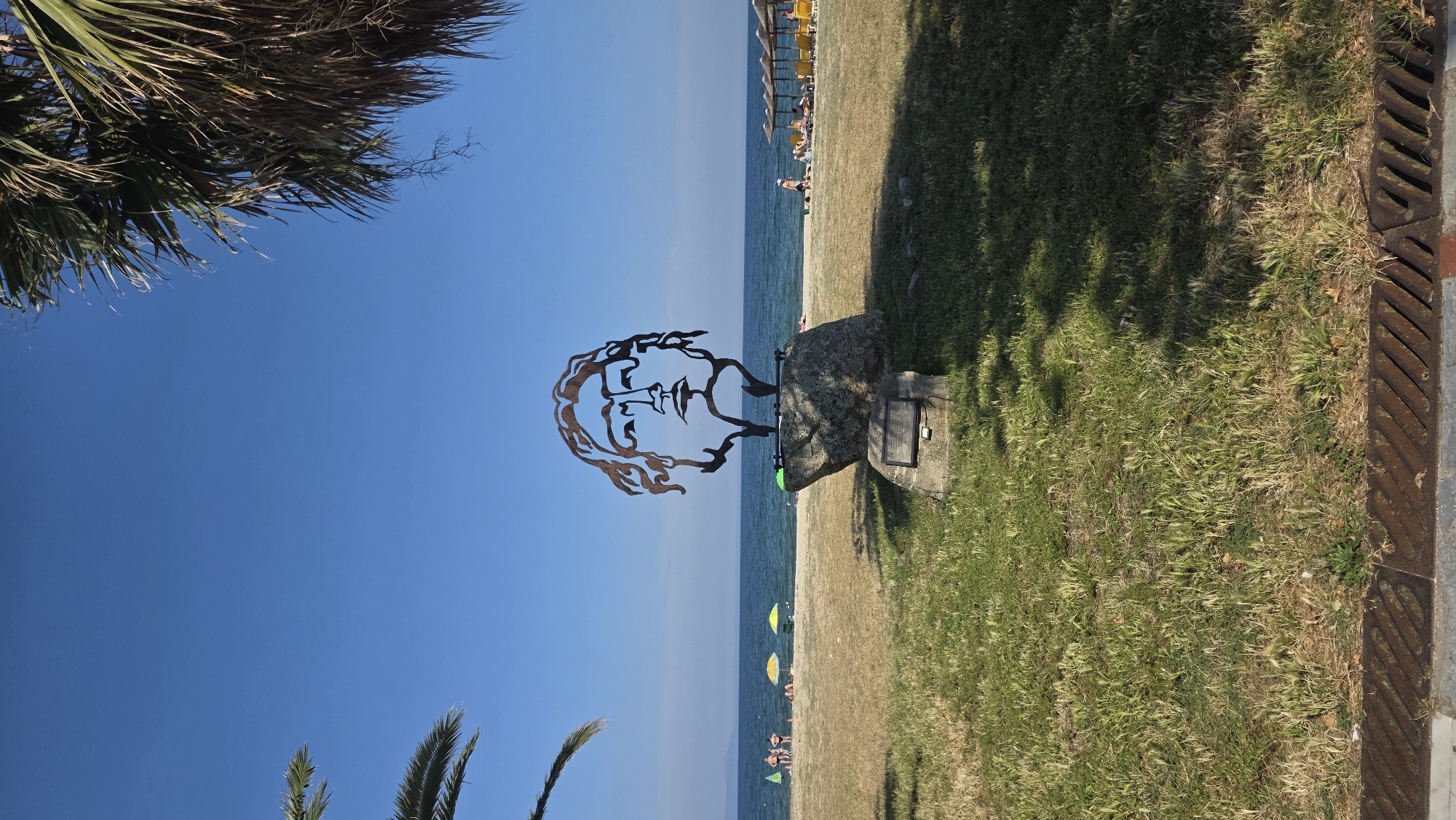 This image depicts a metal sculpture of a human face, rendered in an outlined style, situated on a grassy area adjacent to a beach in Asprovalta, Greece. The sculpture, appearing reddish-brown, is mounted on a large, irregular natural stone boulder, which in turn rests on a smaller, rectangular stone base. A dark, rectangular plaque and a small, black device (possibly a solar light or panel) are attached to the lower stone base.
Behind the sculpture and the grassy strip, a sandy beach extends towards a calm, light blue sea. On the left side of the beach, a small group of approximately four individuals is visible near the waterline, with two yellow and one green beach umbrellas positioned further back on the sand. On the right side of the beach, several individuals are present, some seated on yellow sun loungers beneath a series of thatched umbrellas.
The upper portion of the image is dominated by a clear, unclouded blue sky. Portions of green palm fronds are visible in the upper left and upper right corners, framing the sky. The immediate foreground consists of a patch of green and dry grass, featuring a rusted metal drainage grate and a concrete curb at the bottom edge of the frame. Strong sunlight casts distinct shadows across the grass in the right foreground.