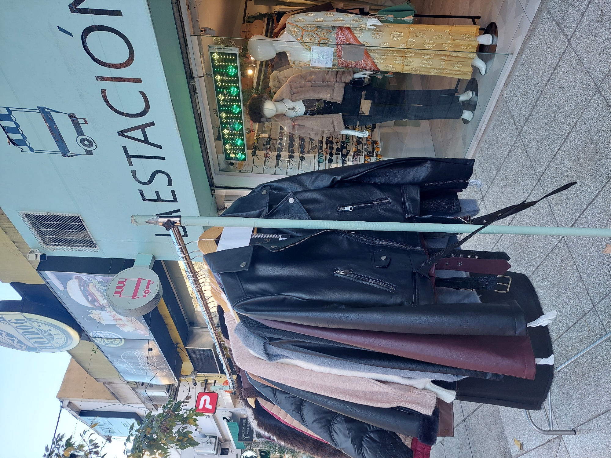 The image shows a sidewalk clothing rack filled with black leather jackets and other garments in front of a store in Buenos Aires, Argentina. The store is called "La Estación." Mannequins are displayed in the store window, wearing clothes and accessories. Street signage for a "Hotdog" restaurant and a logo with the letter "U" are also visible. The background shows a typical urban scene with buildings, trees, and sidewalks.