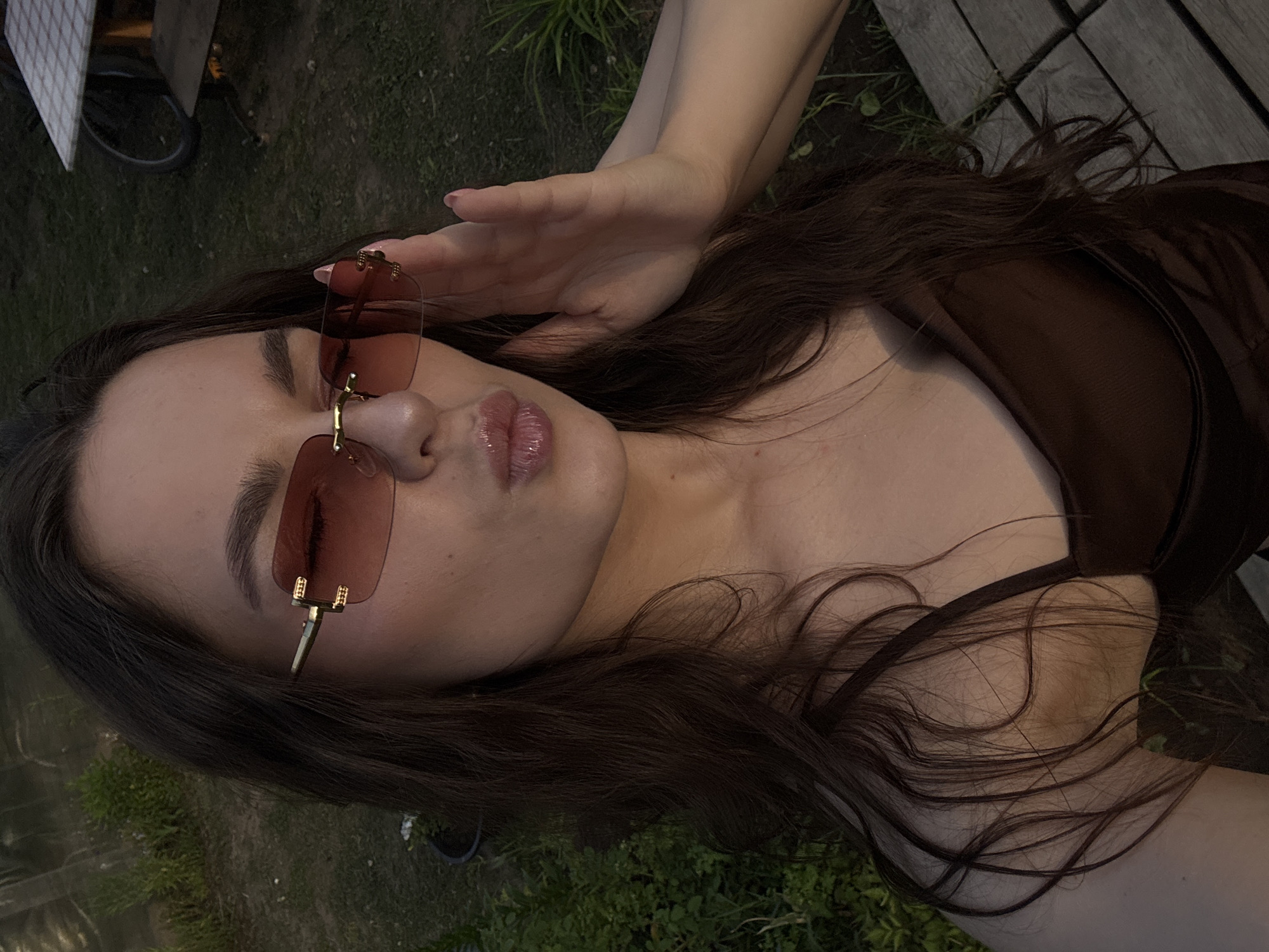A young woman with long, dark brown hair and wearing oversized, rimless sunglasses with rose-tinted lenses is taking a selfie outdoors. She is wearing a dark brown, silky dress with thin straps. Her lips are slightly pursed, and her right hand is raised to the side of her face, near the sunglasses. The background shows greenery and what appears to be a wooden deck or walkway. The lighting suggests it is late afternoon or early evening. The location is identified as Rusalevka, Russia.