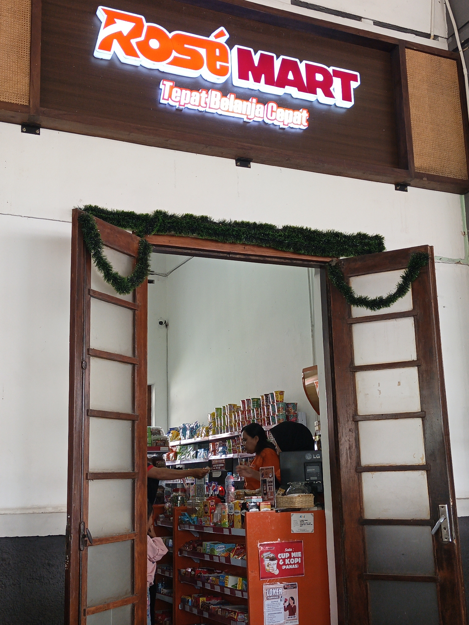 A street-level view of the entrance to a Rosé Mart convenience store in Klaten, Indonesia. The store's name is displayed in large, illuminated red letters above the entrance. Below the main sign, a smaller sign reads "Tepat Belanja Cepat" in white lettering. The entrance features two large, wooden doors, one of which is partially open, revealing the interior of the store. Green garland is draped across the top of the doorway and hangs down on either side. Inside, shelves are stocked with various food items and beverages. A female employee in an orange uniform is visible behind the counter, attending to a customer, a young girl in a pink shirt. The store appears to be open for business.