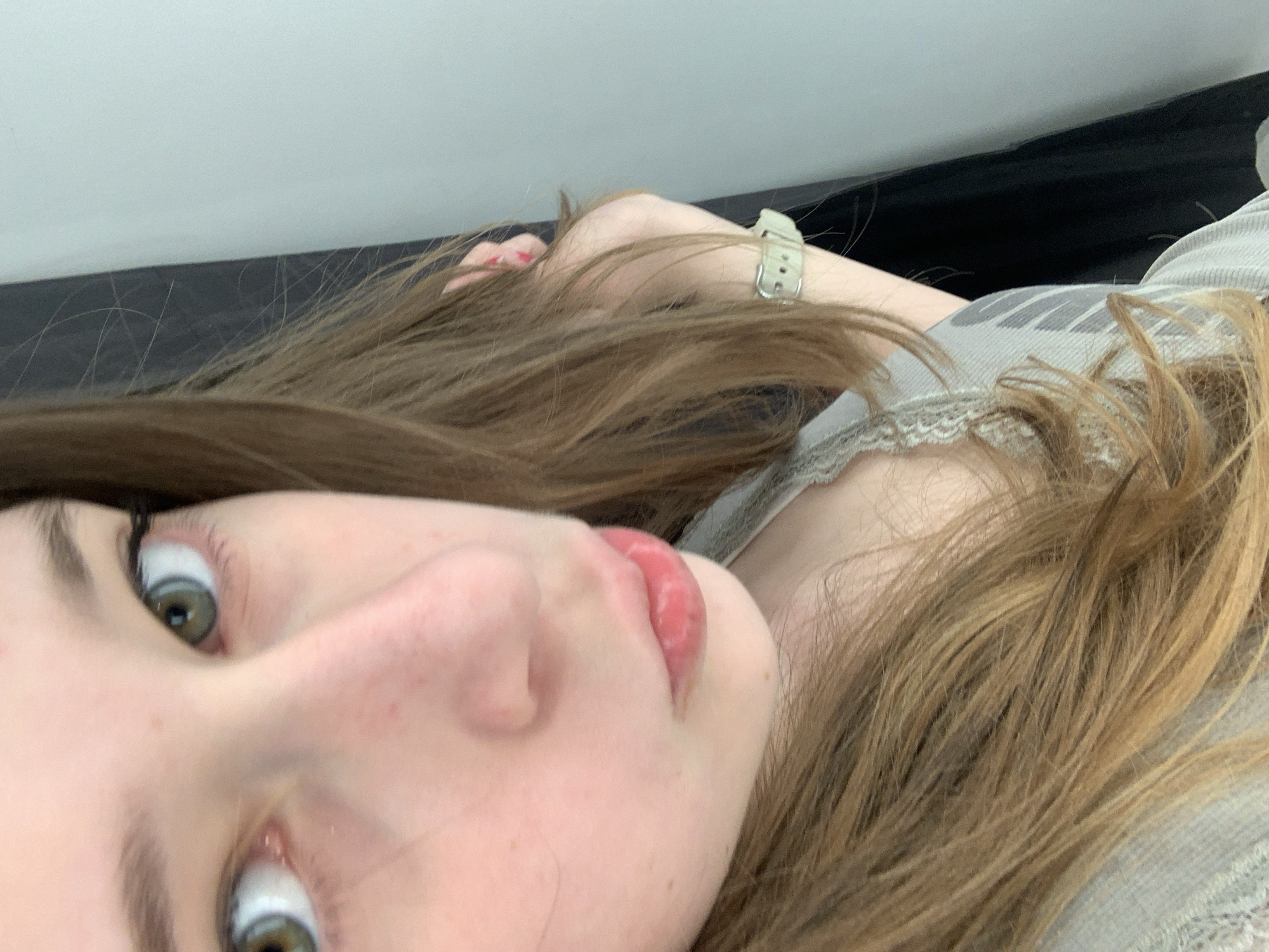 A close-up, angled portrait features a young person, likely female, lying down and looking directly towards the camera. The subject has long, light brown hair spread around their head, prominent green-grey eyes with dark lashes, and slightly parted lips with a glossy, reddish-pink tint. Their expression is direct and engaging.
The person is wearing a light grey, ribbed top adorned with a delicate lace trim along the neckline. A partial graphic or text, showing the letters "NI" in dark grey, is visible on the upper part of the shirt. On their left wrist, a light-colored (possibly beige or off-white) watch with a silver buckle is visible.
The background consists of a dark, uniform surface, likely furniture, and a lighter, solid wall or ceiling above it, indicating an indoor setting. The lighting is soft and even, suggesting an interior environment. The overall impression is casual and personal. The context for this image is Belgrade, Serbia.