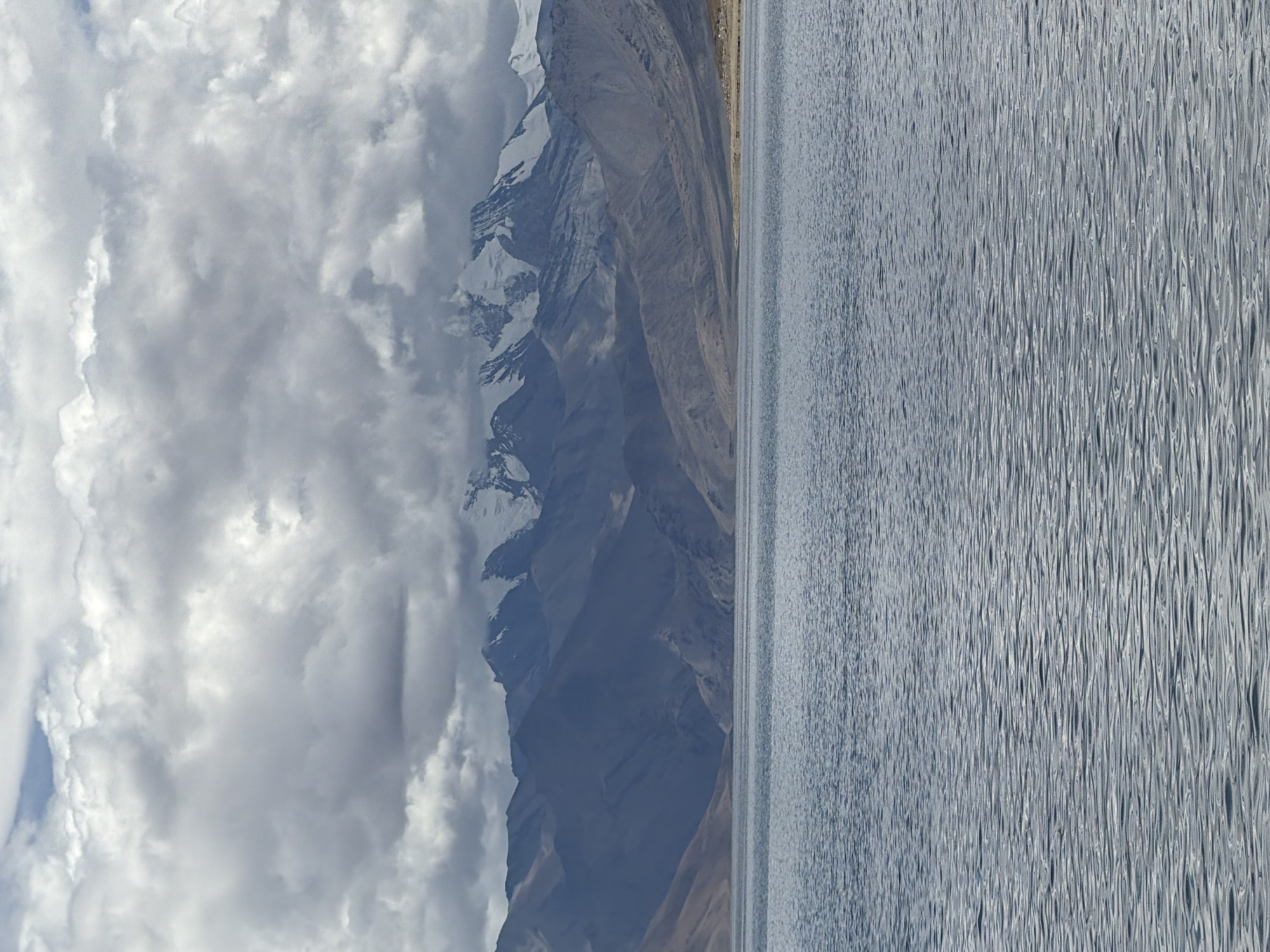 The image depicts a serene natural landscape dominated by a vast body of water in the foreground, reflecting a cloudy sky. In the middle ground, a textured shoreline transitions into a series of rugged, barren mountains. The distant peaks are capped with snow, contrasting with the earth tones of the lower slopes. The sky is filled with dramatic, heavy clouds, suggesting an overcast or impending weather event. The overall impression is one of a remote, mountainous region, possibly a high-altitude lake in India, given the geological features and atmospheric conditions.